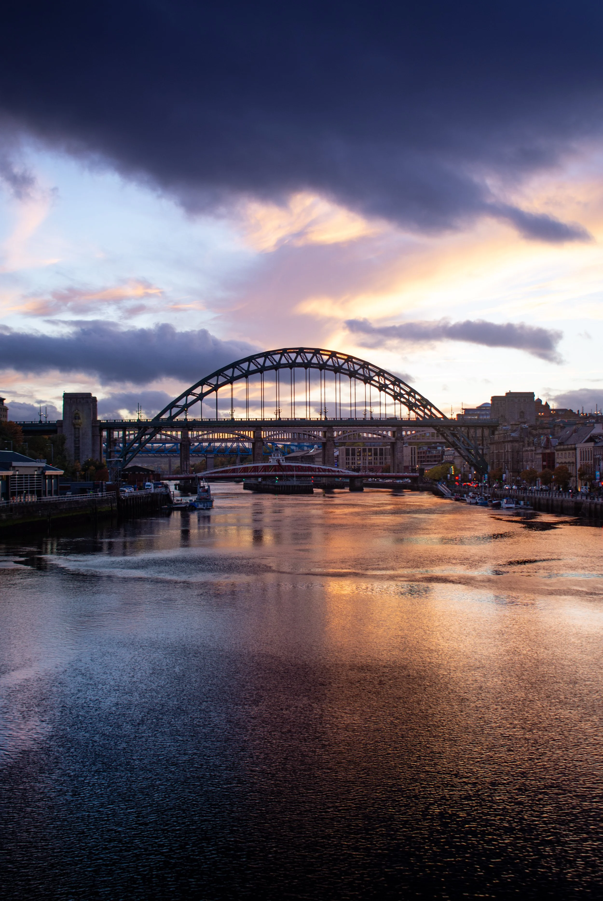 A cityscape view of a bridge over a river at sunset, with buildings along the waterfront and a mostly cloudy sky with some sunlight breaking through.