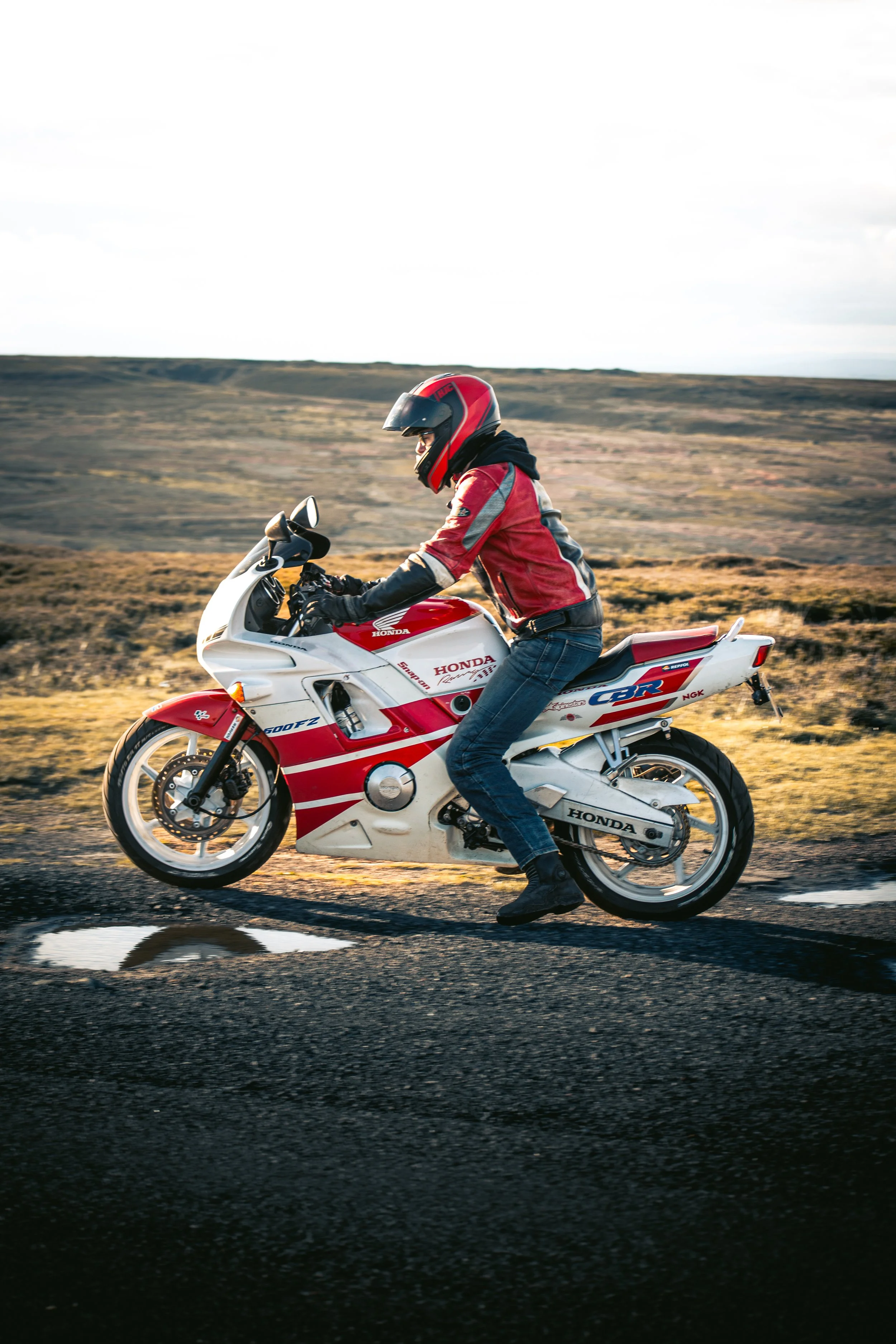 Motorcycle rider wearing a red and black helmet and red jacket riding on a wet road through a rural landscape with fields and hills in the background.
