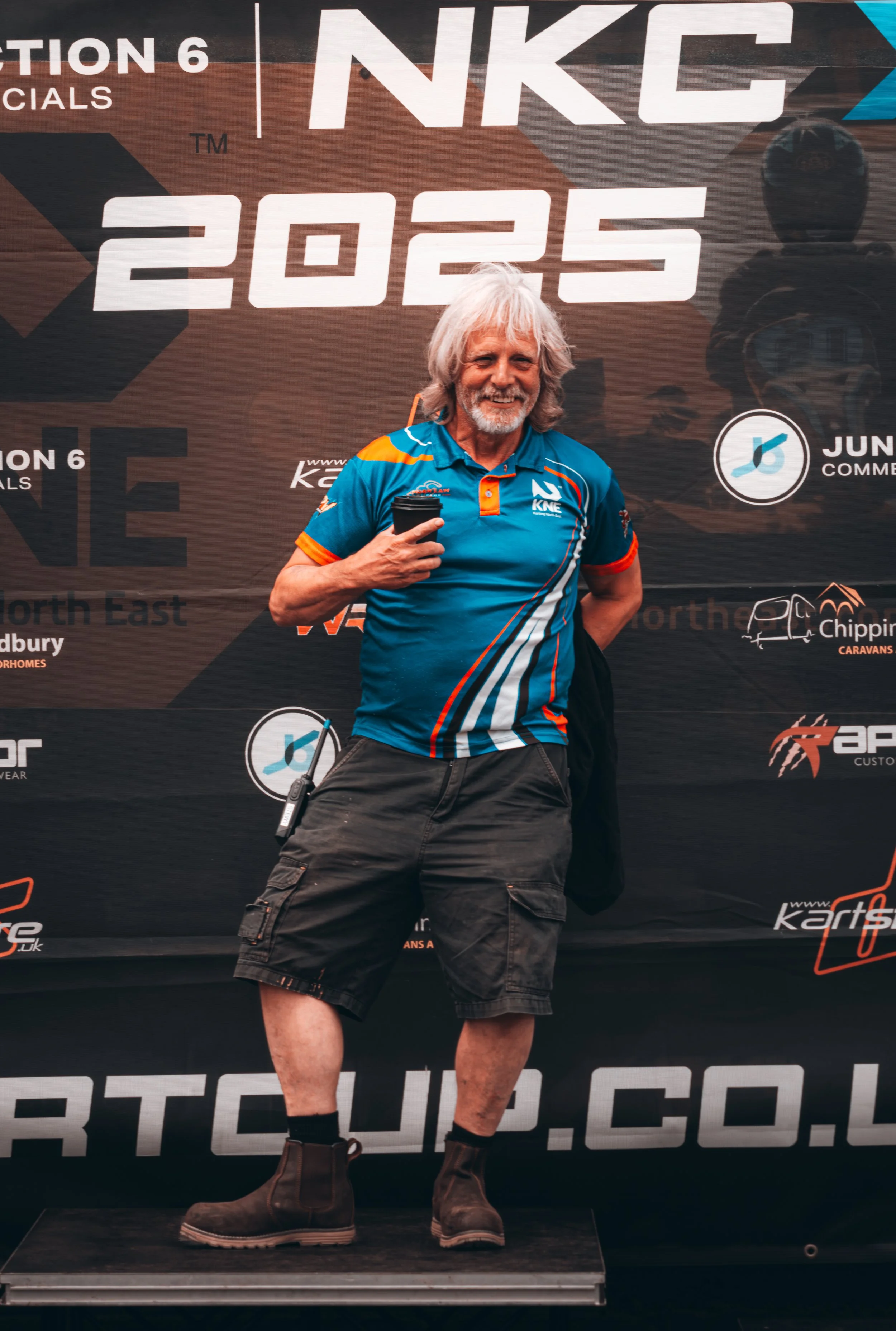 A man with gray hair and beard smiling, wearing a blue racing shirt with orange and white accents, black cargo shorts, and black boots, holding a coffee cup, standing on a podium in front of a black backdrop with logos and large text for the 2025 NKC racing event.