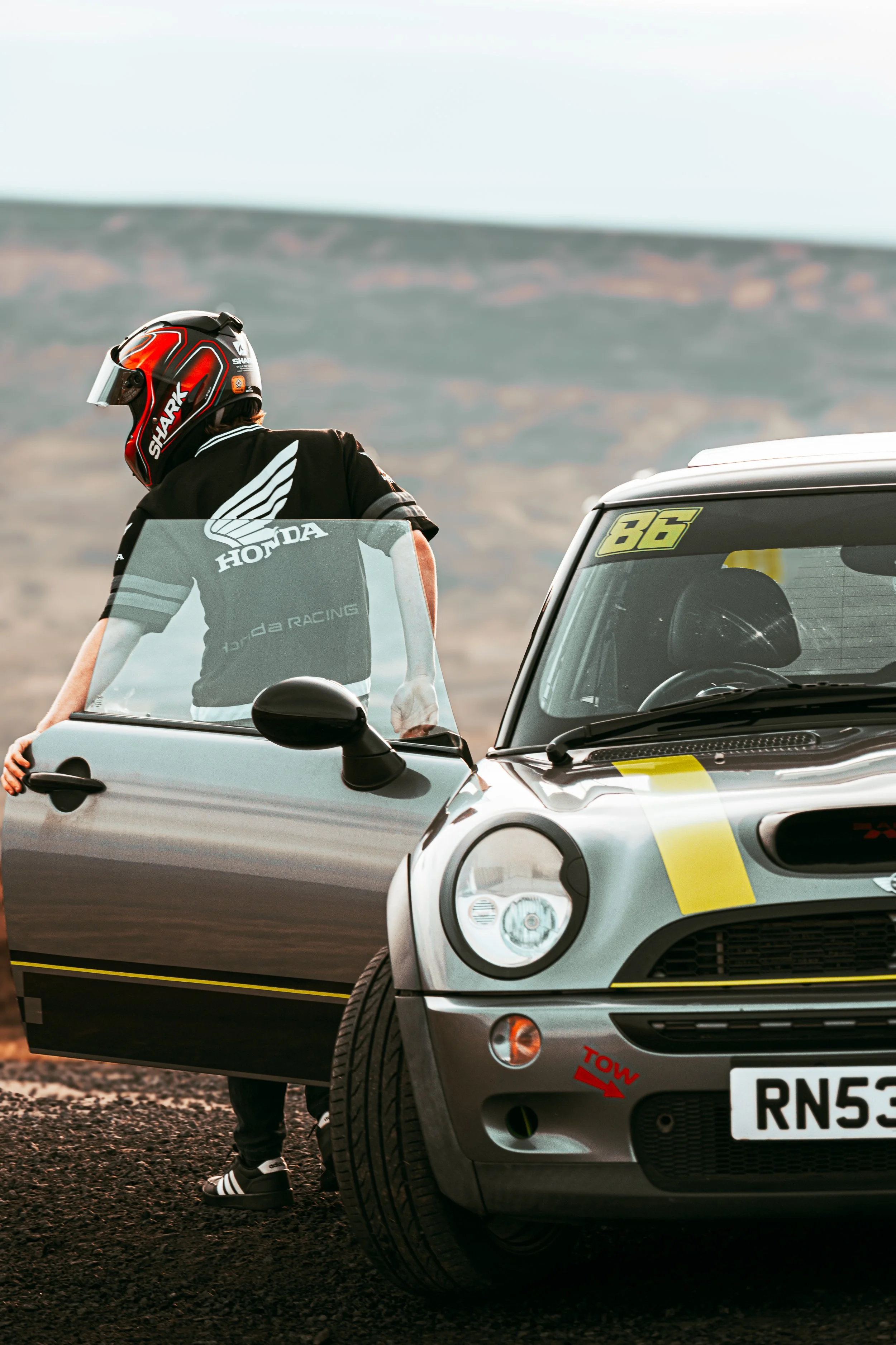 A person wearing a helmet and black Honda racing shirt standing next to a silver Smart car with racing stickers in a desert landscape.