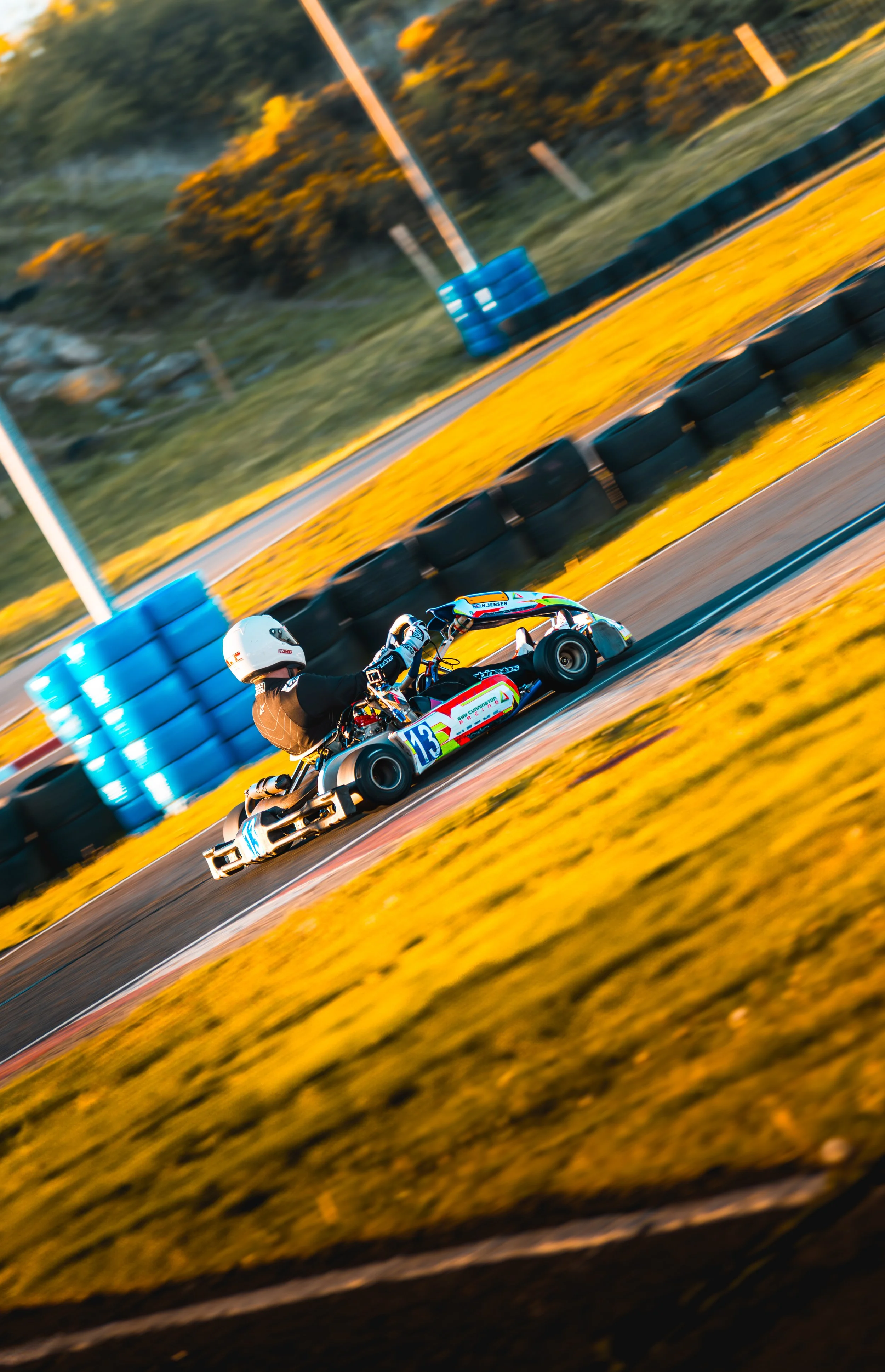 A race car driver in a helmet and racing suit driving a go-kart on a race track with yellow and black tire barriers and a grassy hill in the background.