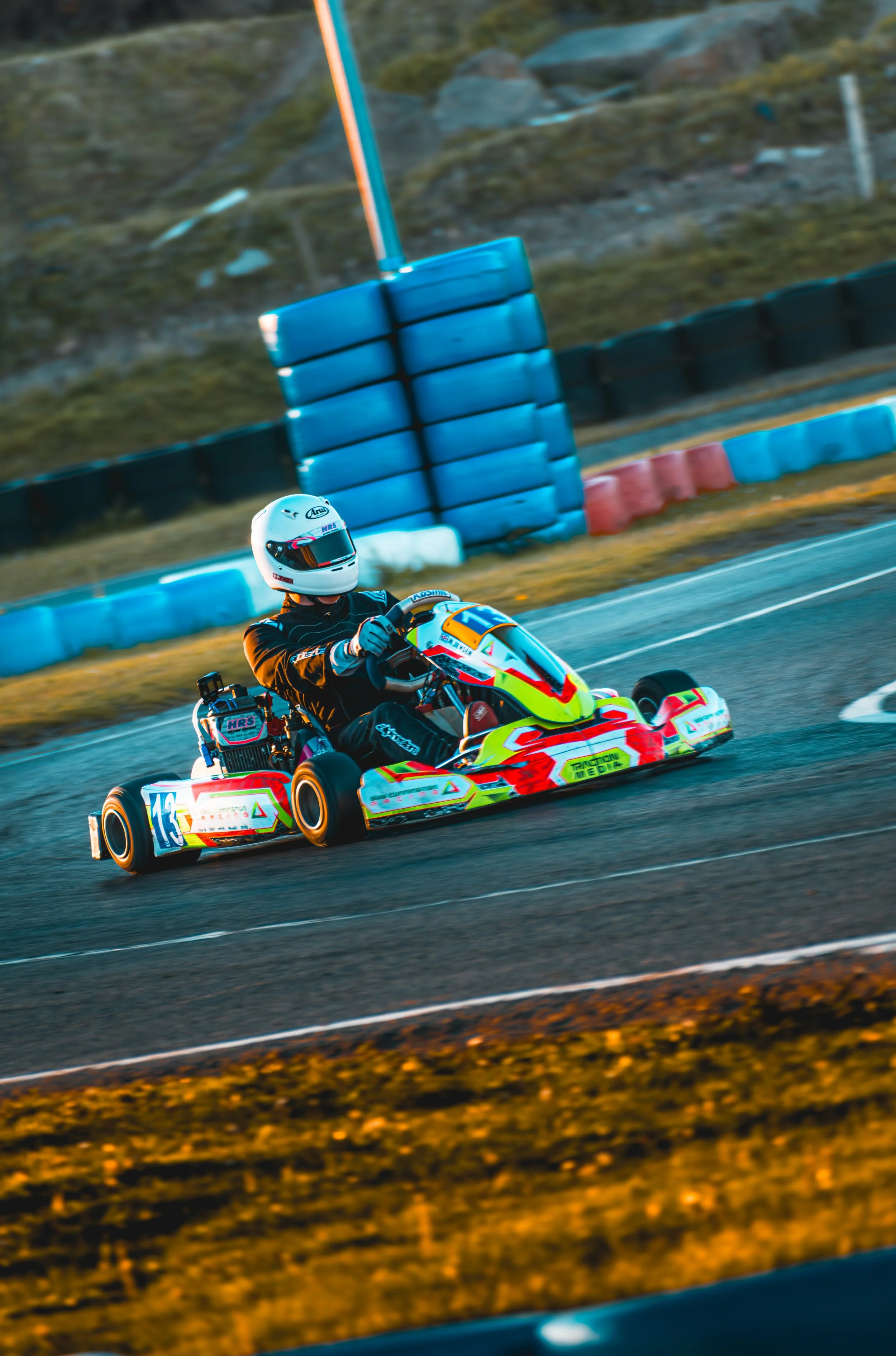 A go-kart driver wearing a white helmet and a black racing suit, navigating a turn on a race track with blue, black, and orange barriers.