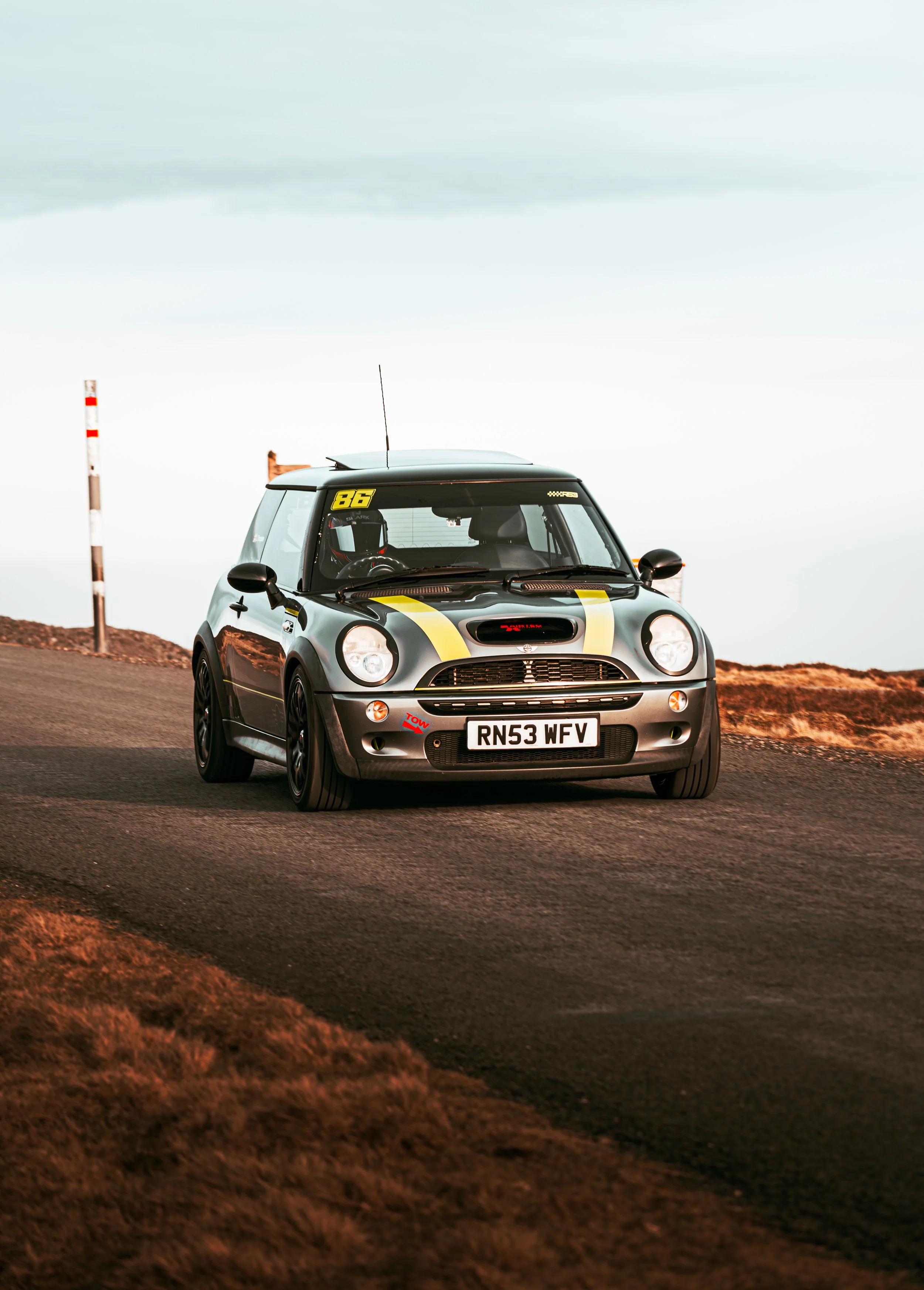 A small black and yellow race car with the number 86 on the windshield, parked on a road with a dirt embankment on the side, under a cloudy sky.