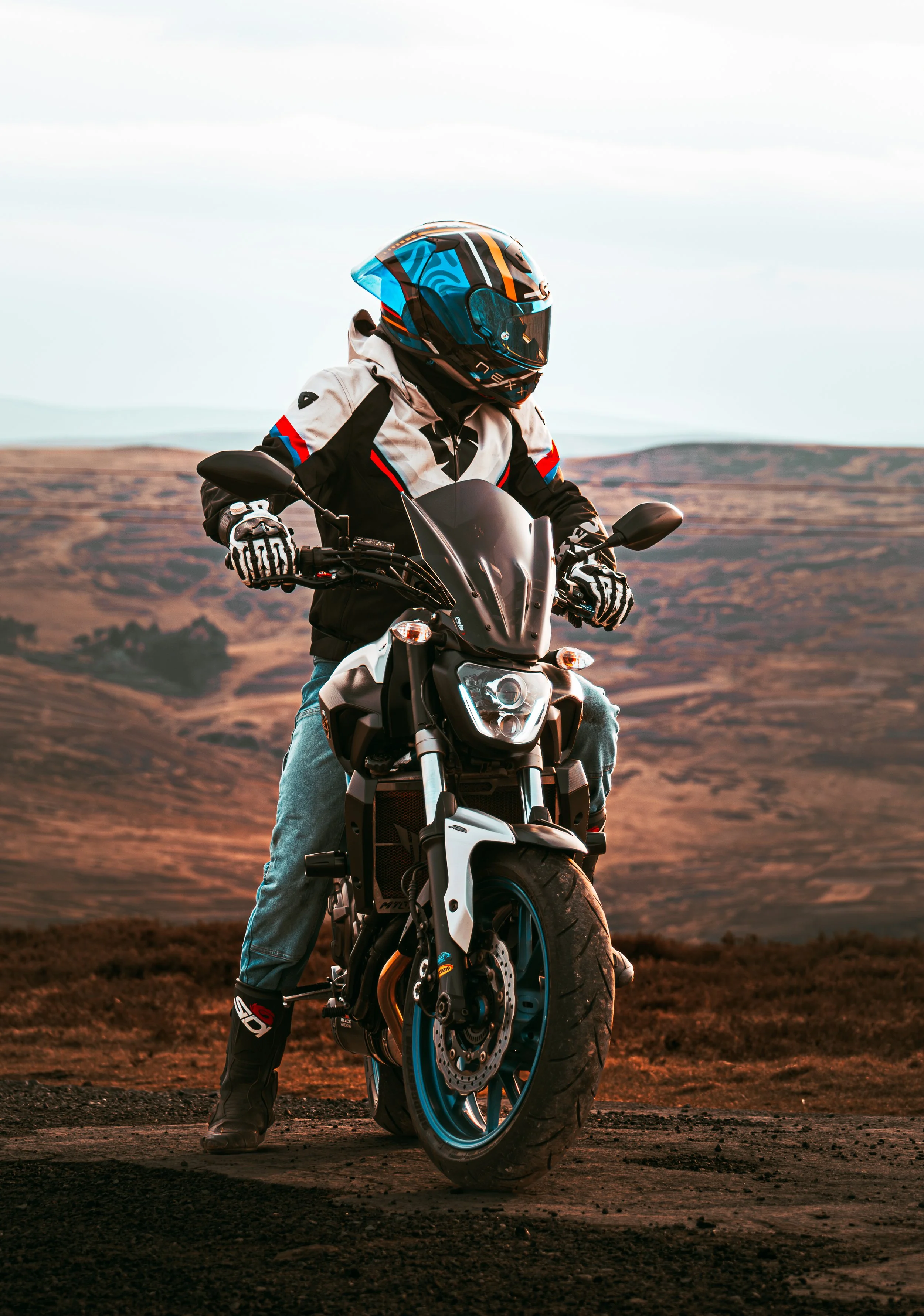 Motorcycle rider wearing a colorful helmet and riding gear, standing on a dirt road with open landscape and hills in the background during sunset.