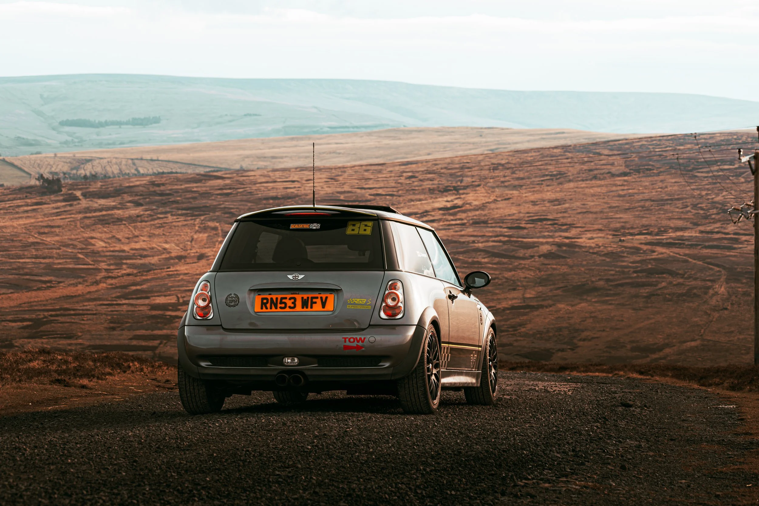 A silver Mini Cooper car with racing stickers and a UK license plate, parked on a dirt road in a rural landscape with rolling hills in the background.