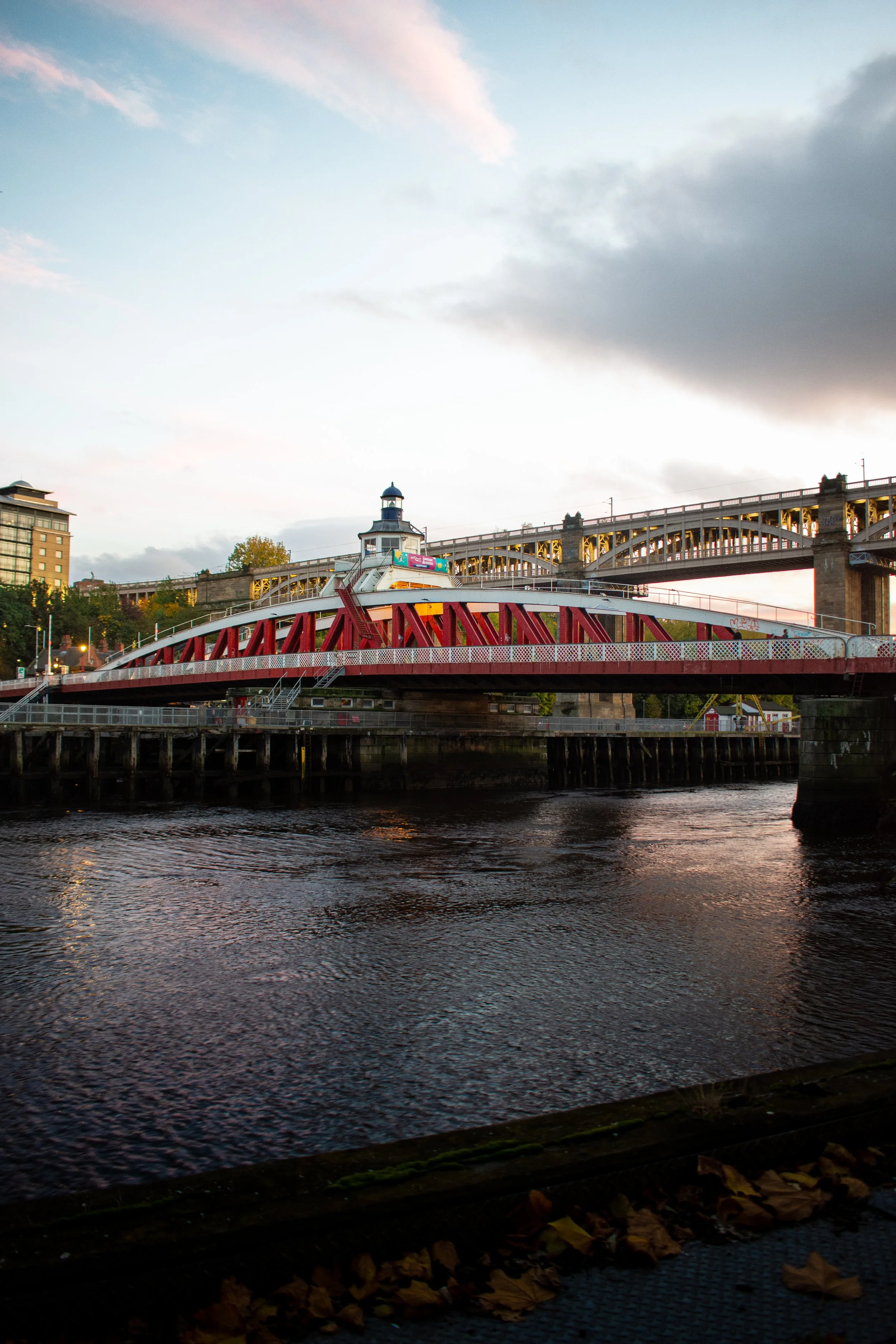 A waterfront scene during sunset with a bridge and a lighthouse-like structure, some leaves on the ground, and buildings in the background.