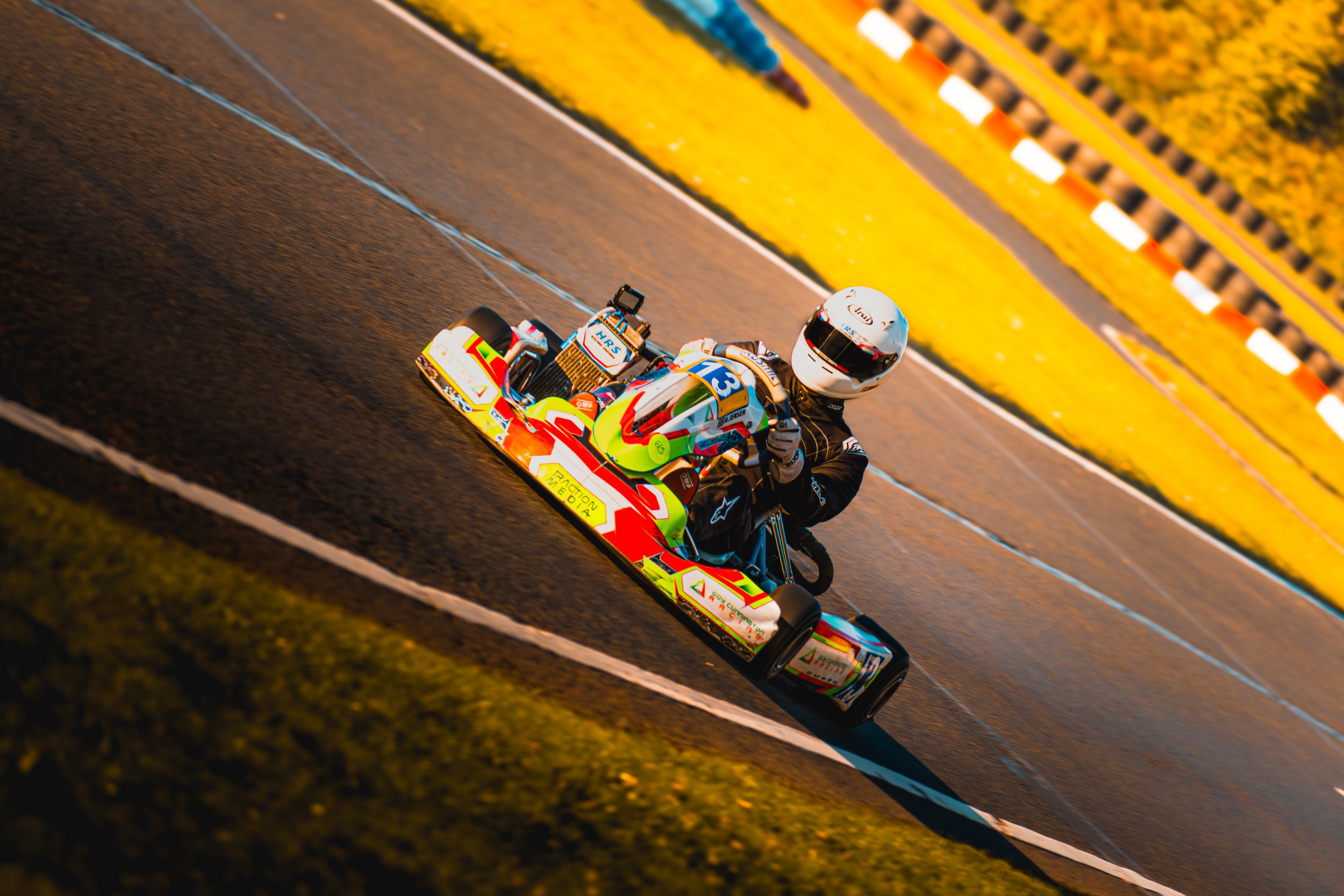A race car with the number 13 on it, driven by a person wearing a white helmet, on a paved race track during autumn with yellow trees in the background.