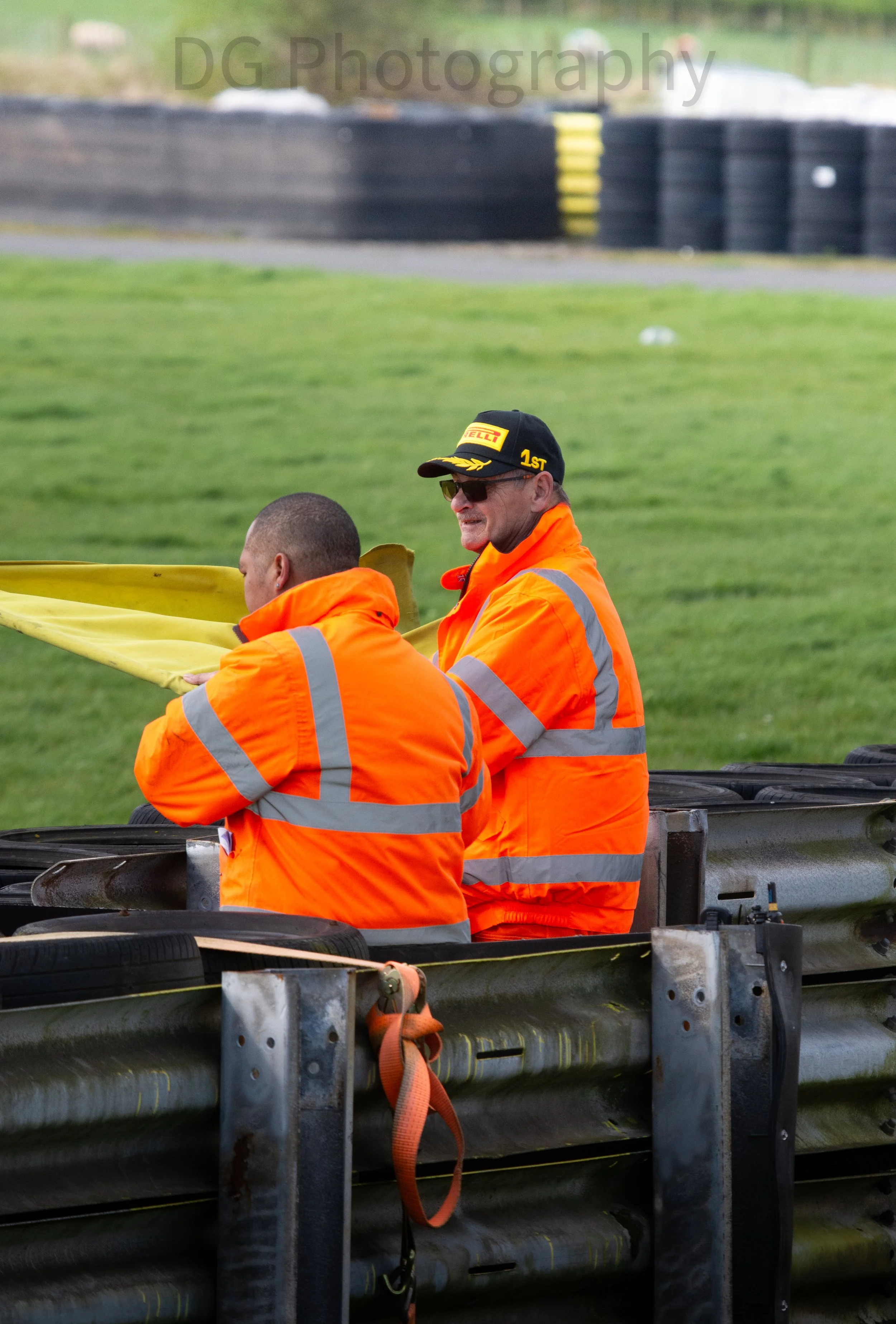 Two men in orange safety jackets sitting on a boat platform with tires attached, engaging in an activity near a grassy area. One man is wearing a black cap with yellow accents and sunglasses, the other has a shaved head.