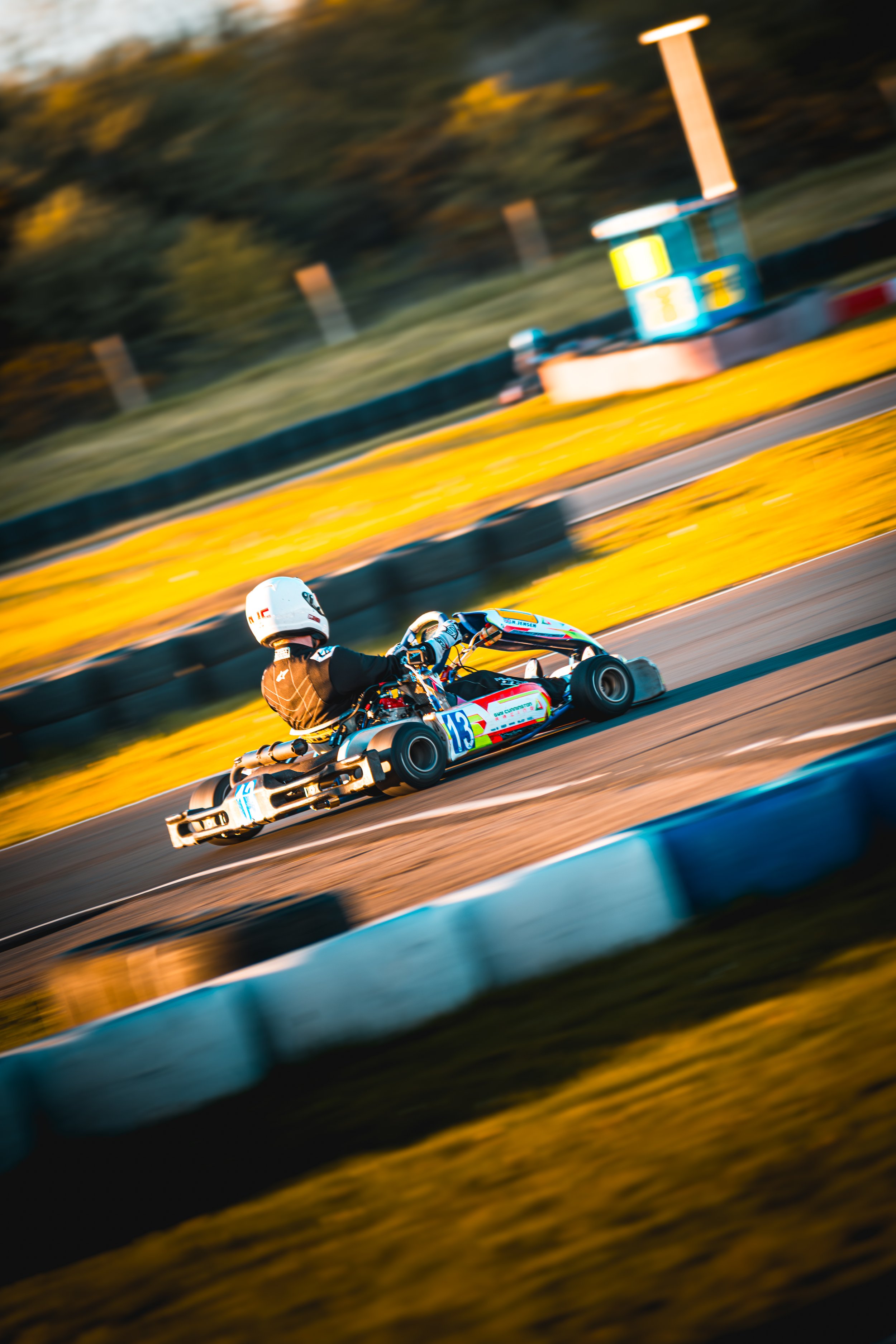 A person racing a go-kart on an outdoor track with yellow barriers, wearing a white helmet and black racing suit. The scene is captured mid-movement with a blurred background.