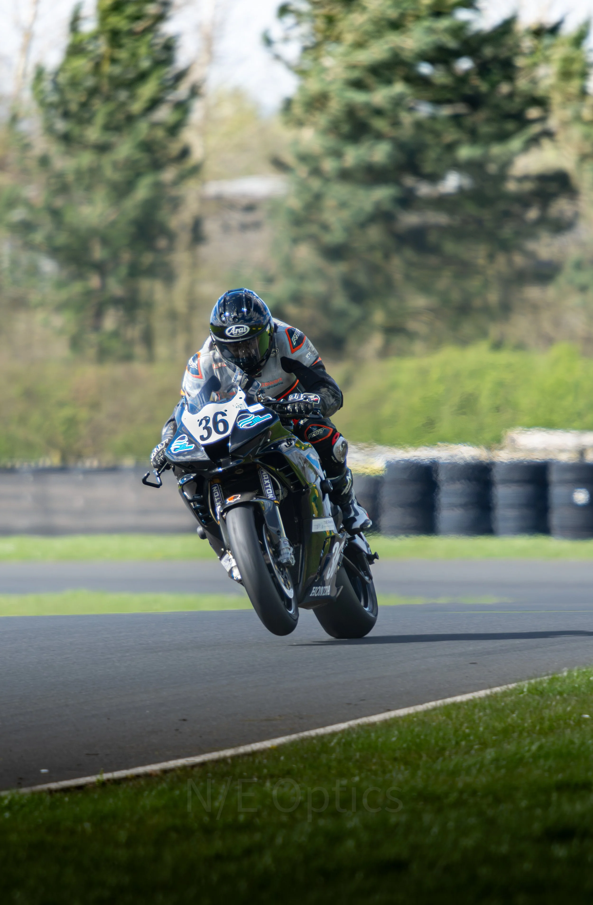A motorcyclist wearing a helmet and racing suit leaning into a curve on a racetrack, with trees blurred in the background.
