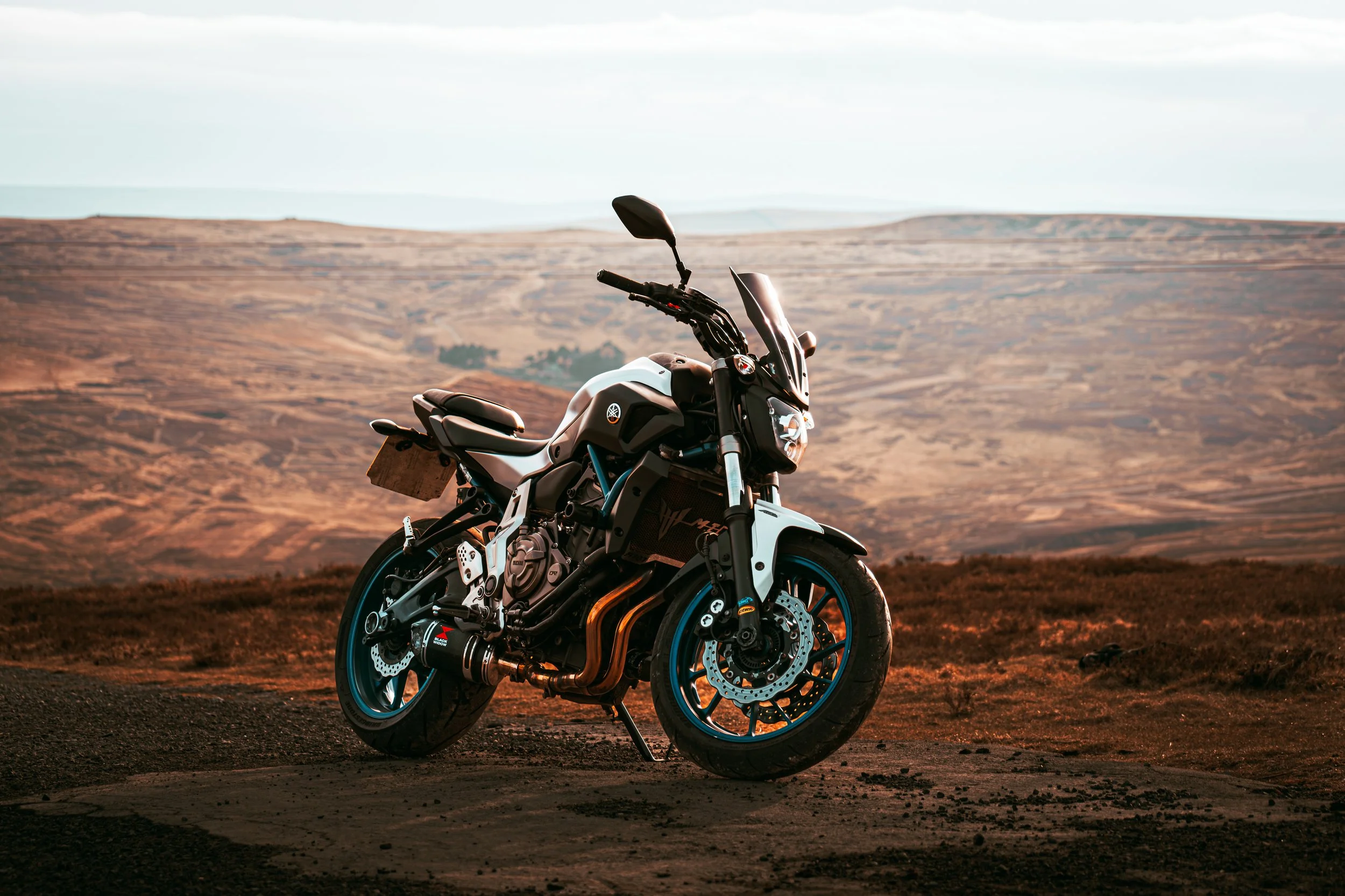 A black and white motorcycle parked on a dirt road in a desert landscape with rolling hills and cloudy sky in the background.