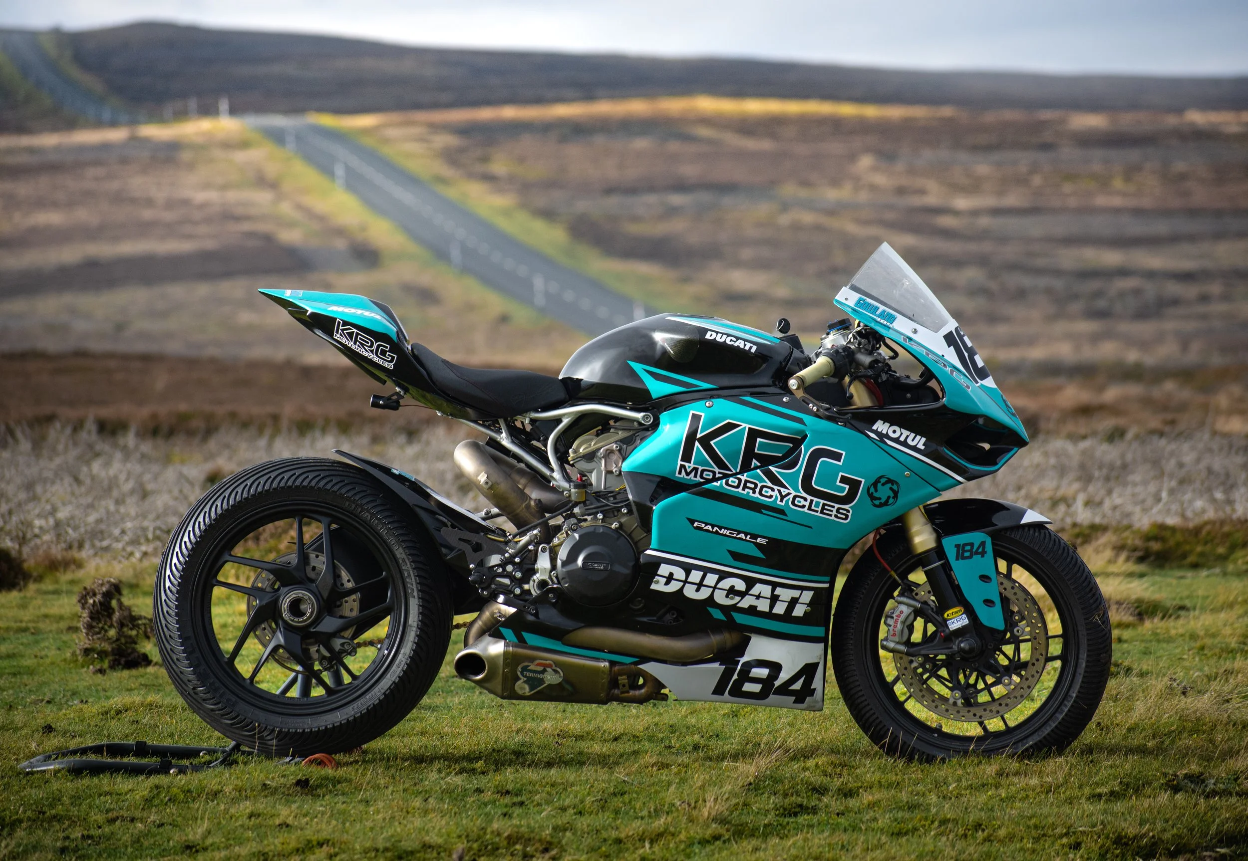 A blue and black Ducati racing motorcycle parked on a grassy field with a winding road and hills in the background.