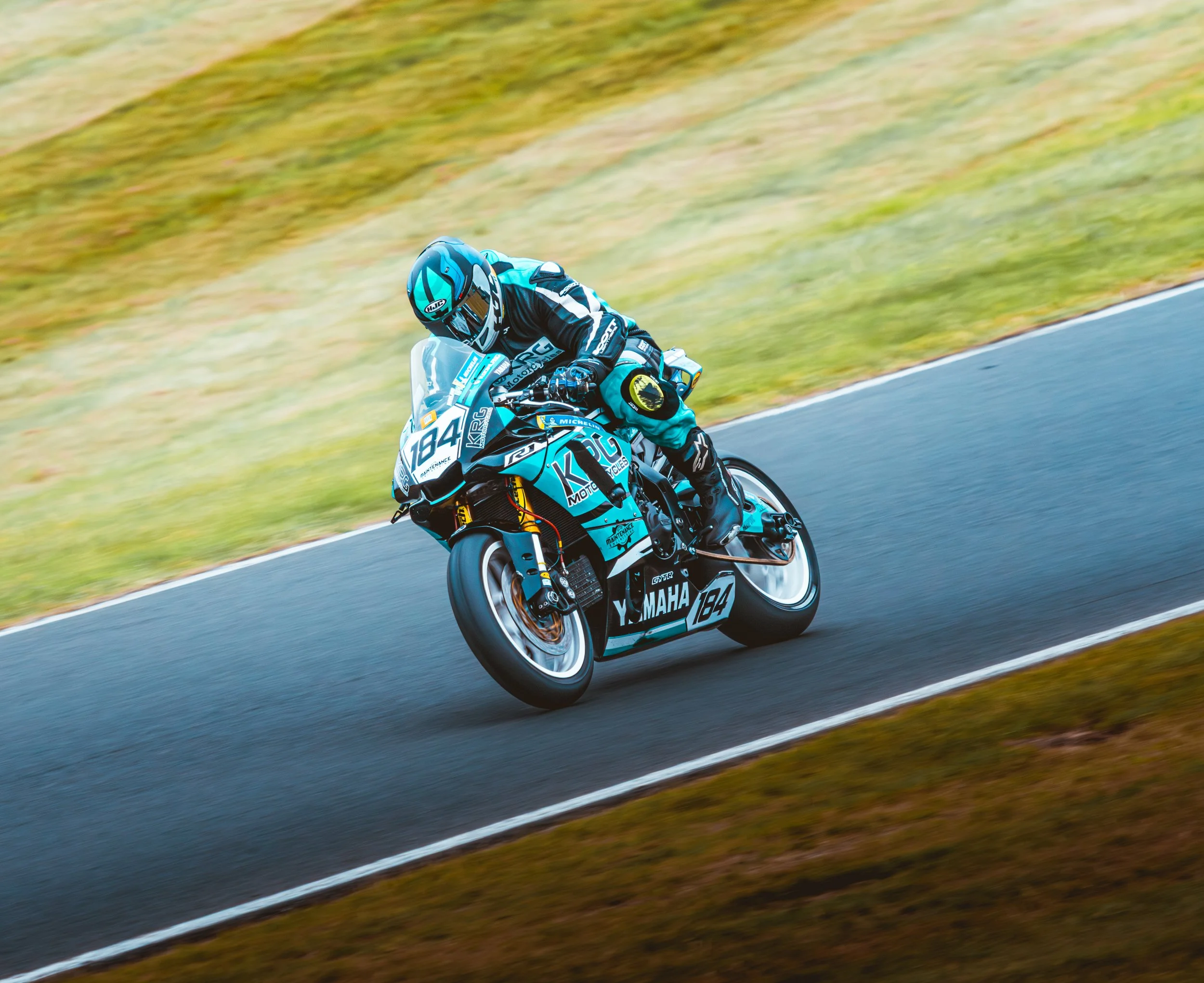 Motorcycle racer wearing a blue helmet and racing suit, leaning into a turn on a racetrack surrounded by grassy area.