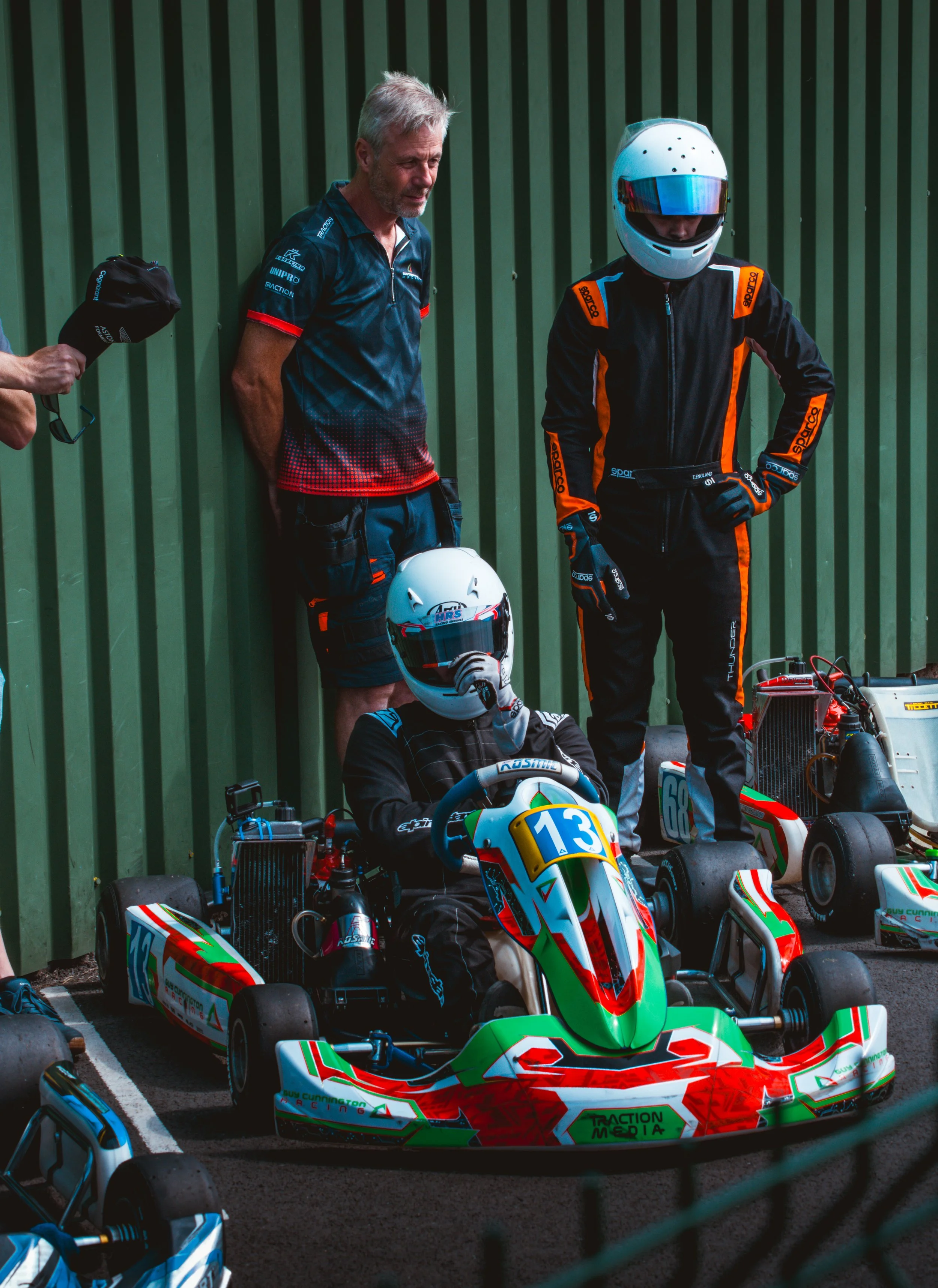 A group of go-kart racers and their mechanic or coach standing beside green and red go-karts, with one racer wearing a racing helmet and suit, and others with helmets off, in a pit area with a green corrugated wall in the background.