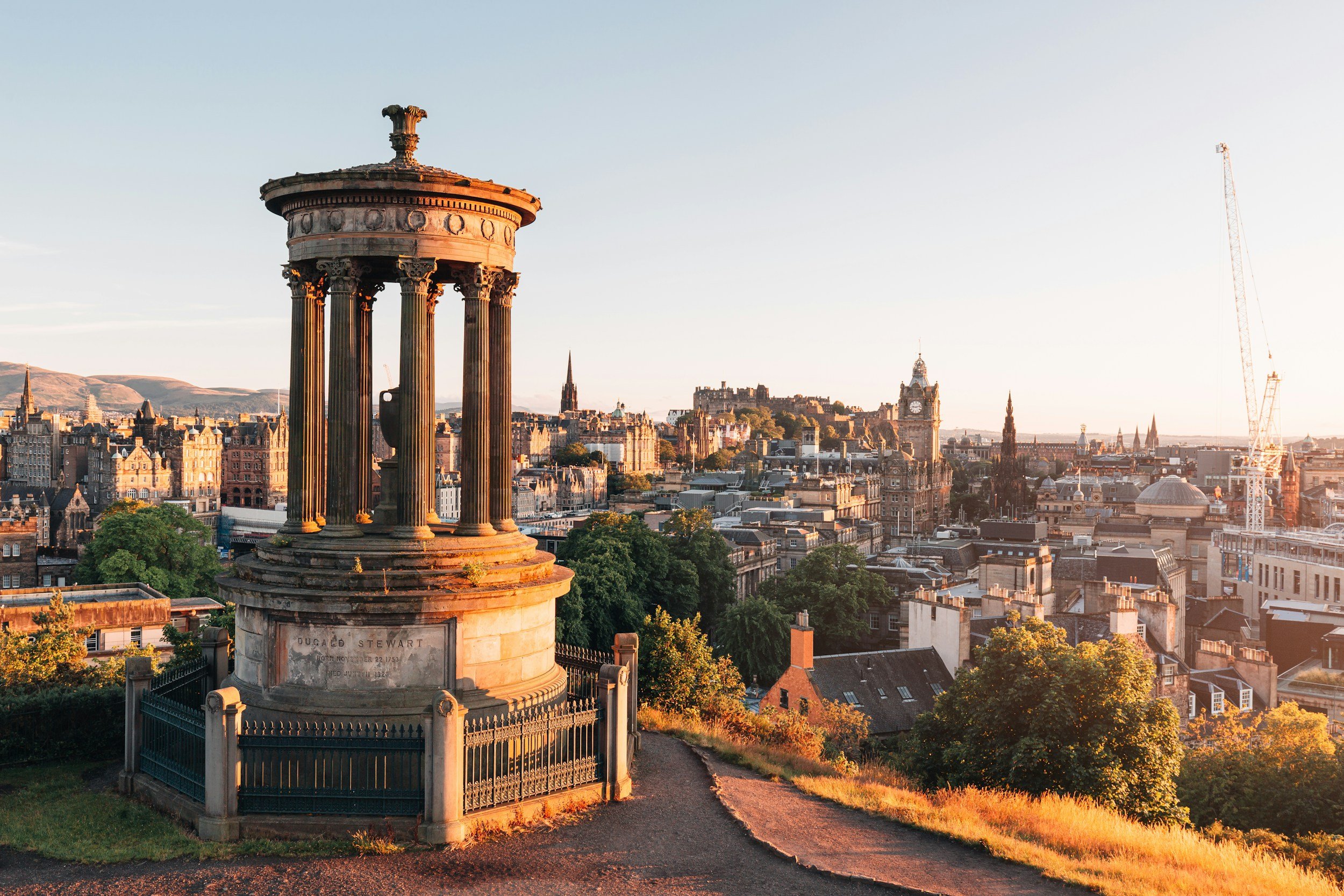 A stone monument with columns and a statue on top on a hill overlooking a cityscape of Edinburgh, Scotland, at sunset. Prominent landmarks include the Edinburgh Castle and Big Ben.