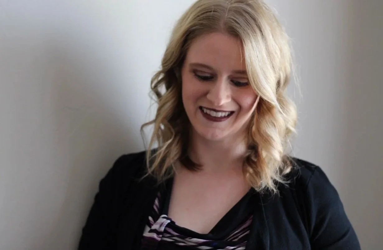A young woman with blonde, wavy hair is smiling and looking down, wearing a black jacket over a patterned top, against a plain wall background.