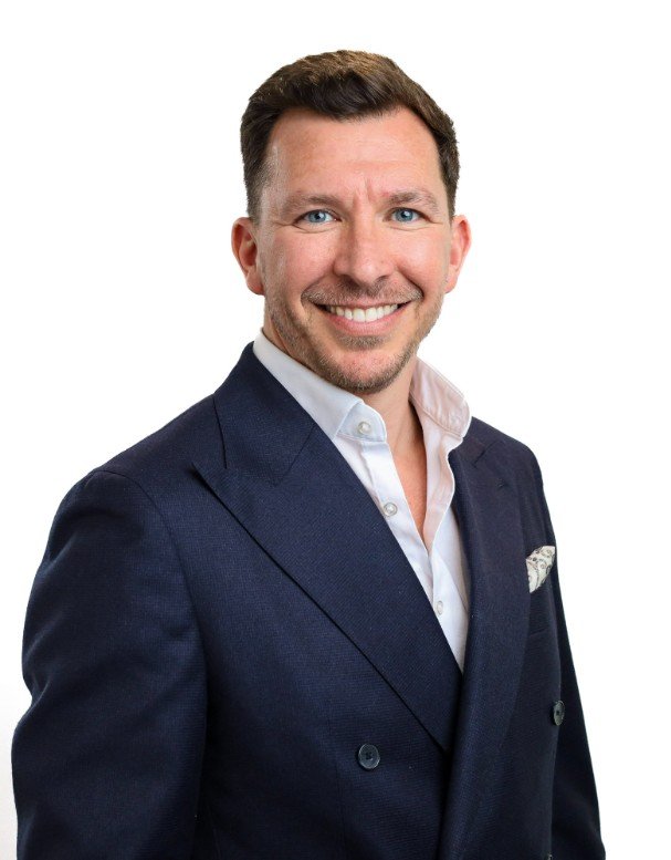 A smiling man with short brown hair, light facial hair, wearing a navy blue blazer over a white dress shirt, and a patterned pocket square, against a plain white background.