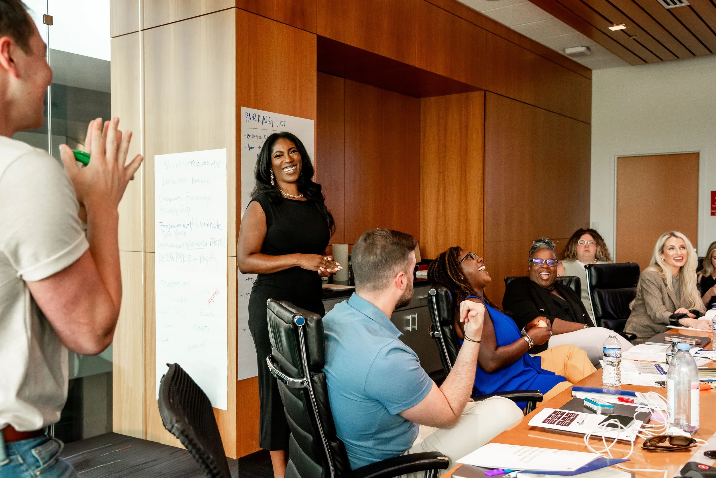 A diverse group of people attending a meeting or workshop in a conference room. A woman in a black dress is smiling and standing, while others are seated around a table with papers, water bottles, and electronic devices.
