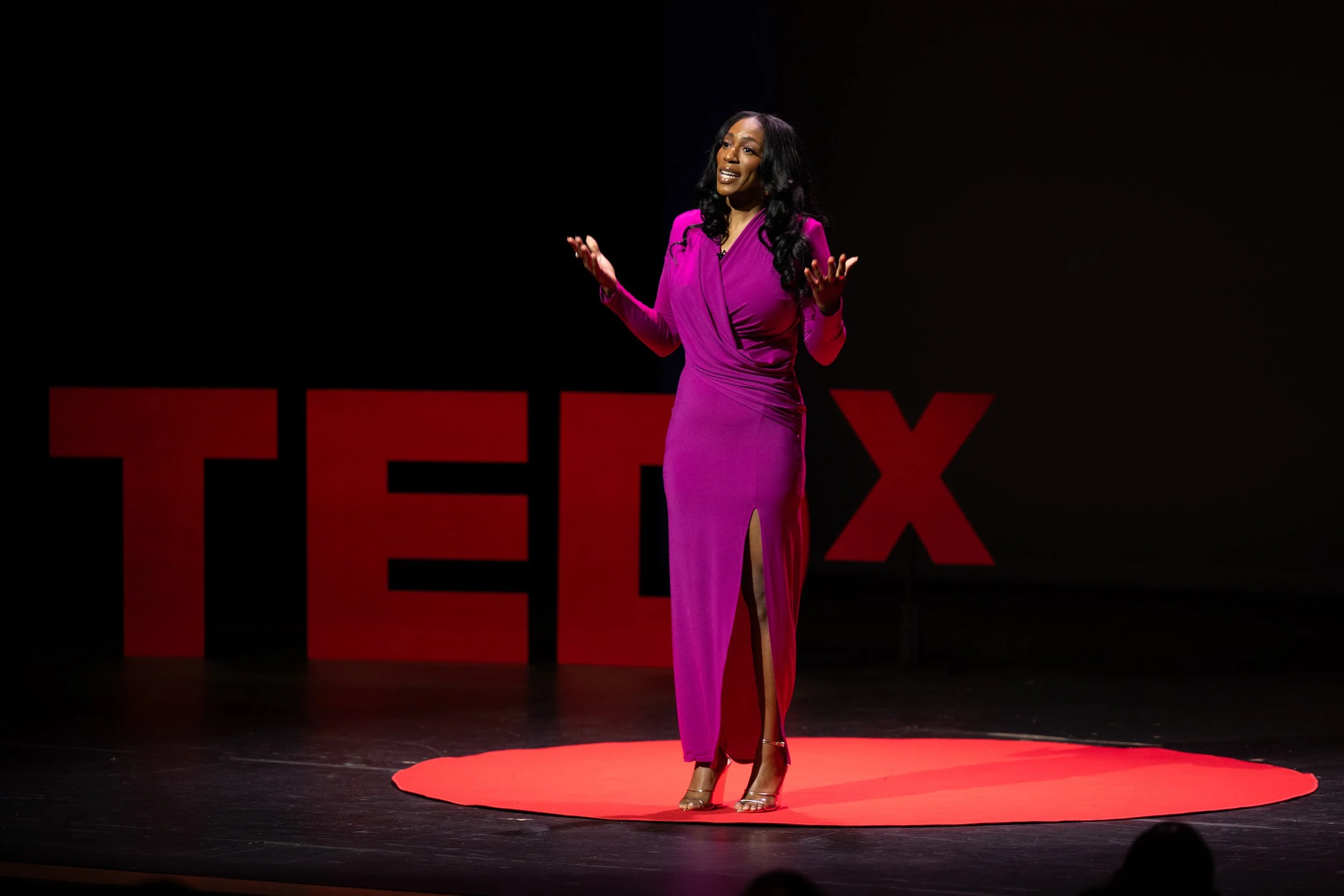 A woman in a long, magenta dress giving a presentation on a stage with a dark background and large red and black TEDx logos.