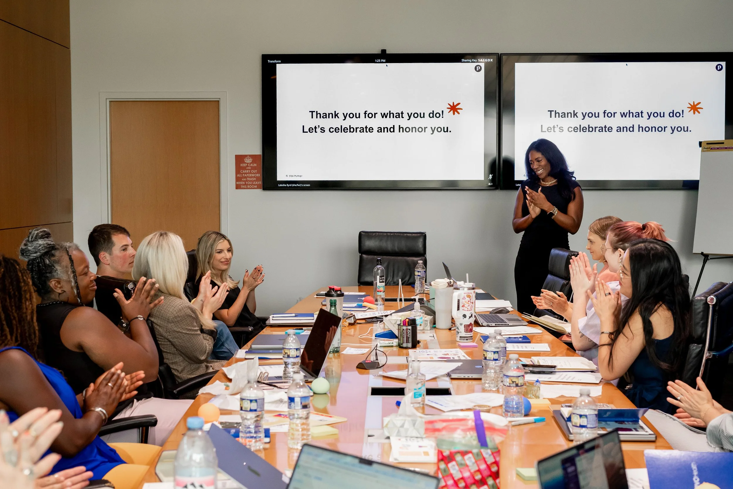 A diverse group of people sitting around a conference table, clapping and smiling as a woman stands and speaks, with two large screens behind her displaying a message of appreciation and celebration.