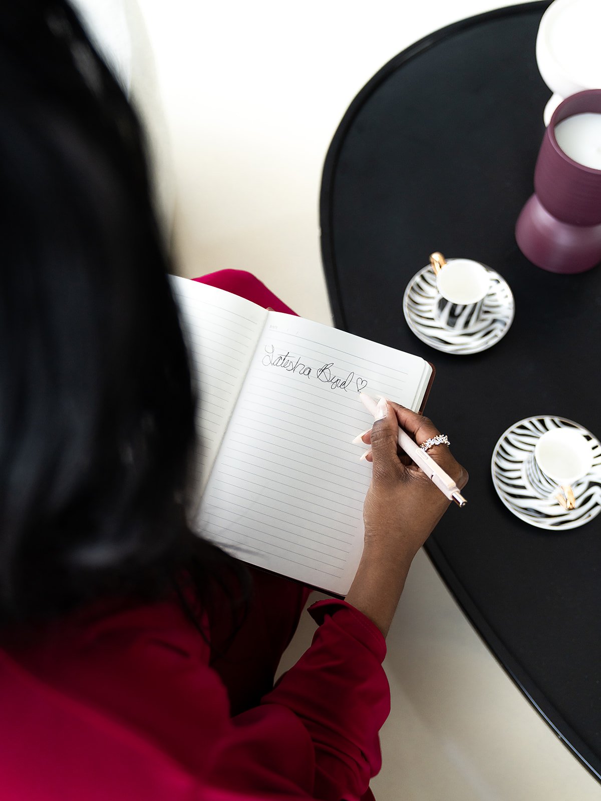 A person writing in a notebook with the words "Latetra Good" and a heart symbol. There are decorative plates and candle holders on the black table.