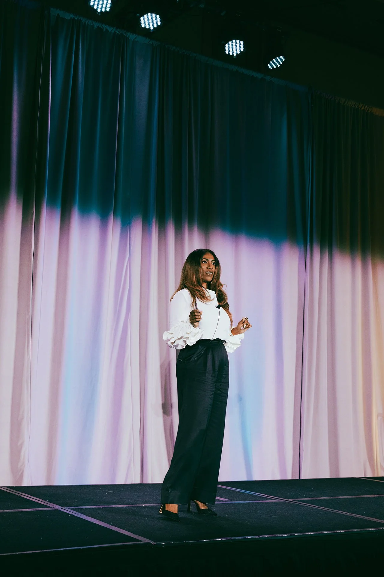 A woman with long, wavy hair stands on a stage with a multi-colored curtain behind her, speaking into a microphone.