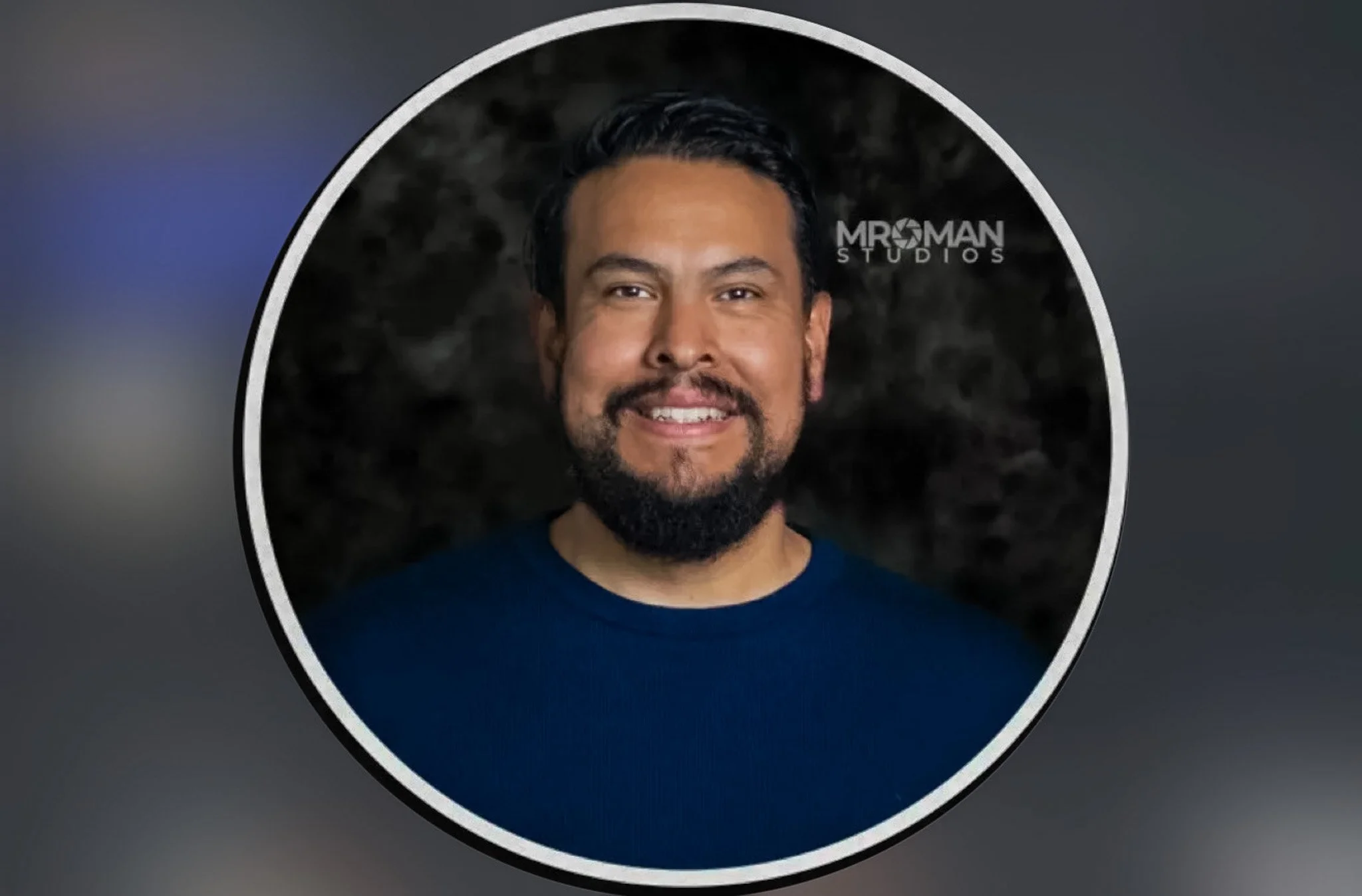 A man with dark hair and a beard smiling, wearing a blue shirt, against a dark patterned background. The picture is framed in a circular border with a white outline. The logo of MRO Man Studios is visible to the right of the man's head.
