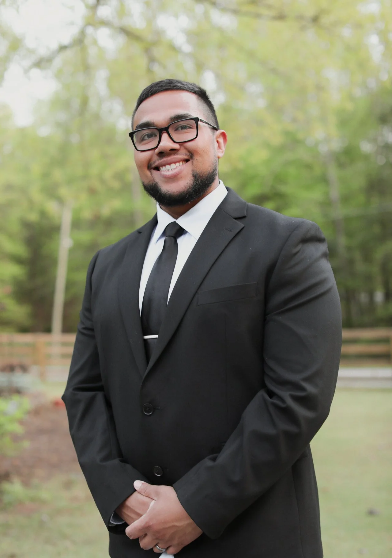 Young man in black suit and tie, wearing glasses, smiling outdoors with green trees in the background.