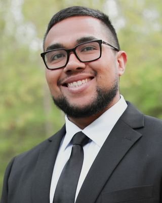 A young man with glasses and a beard, wearing a black suit and tie, smiling outdoors with greenery in the background.