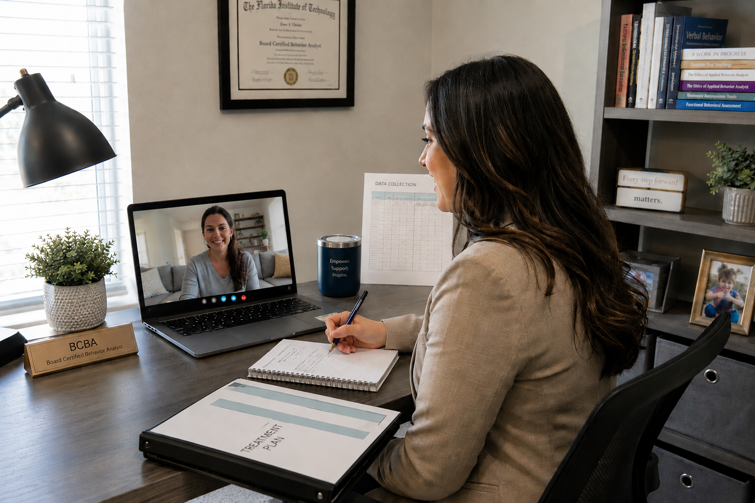 A woman in a beige blazer sitting at a desk, engaging in a video call with another woman on a laptop. The desk has a binder labeled 'Treatment Plan', a notebook, a nameplate reading 'BCBA', a potted plant, and a bottle with a motivational message. Behind her is a bookshelf with books and framed pictures, a certificate on the wall, and a window with blinds.