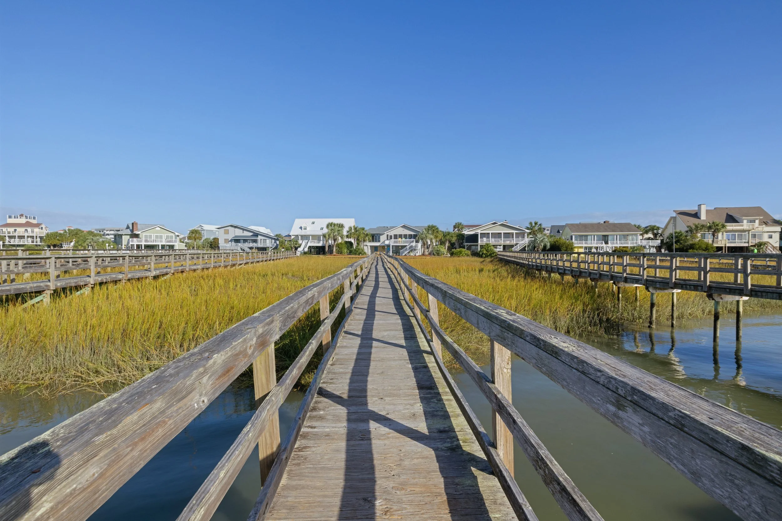 Wooden boardwalk over marshland with houses and palm trees in the background under a blue sky.