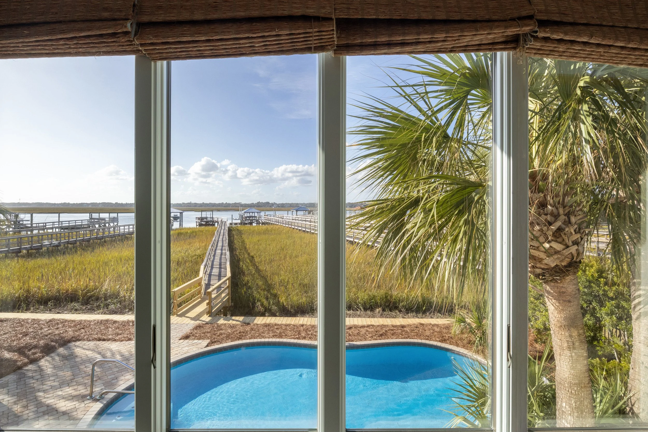 View through glass doors of a backyard swimming pool and a wooden boardwalk leading to a dock over a marsh or river with palm tree on the right and a clear sky with some clouds.