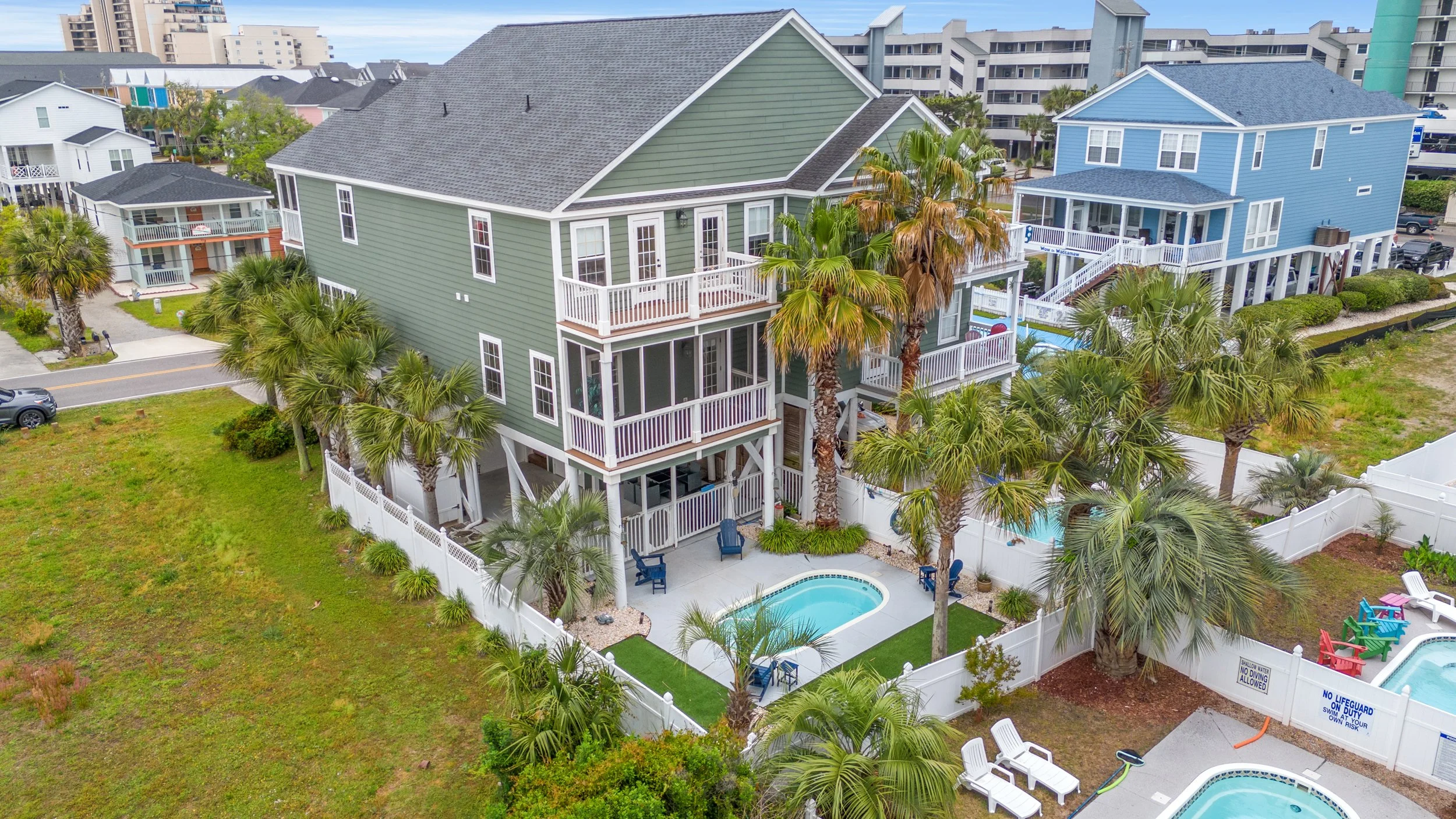 Aerial view of colorful beach houses with balconies, palm trees, swimming pools, and a fenced backyard on a sunny day.