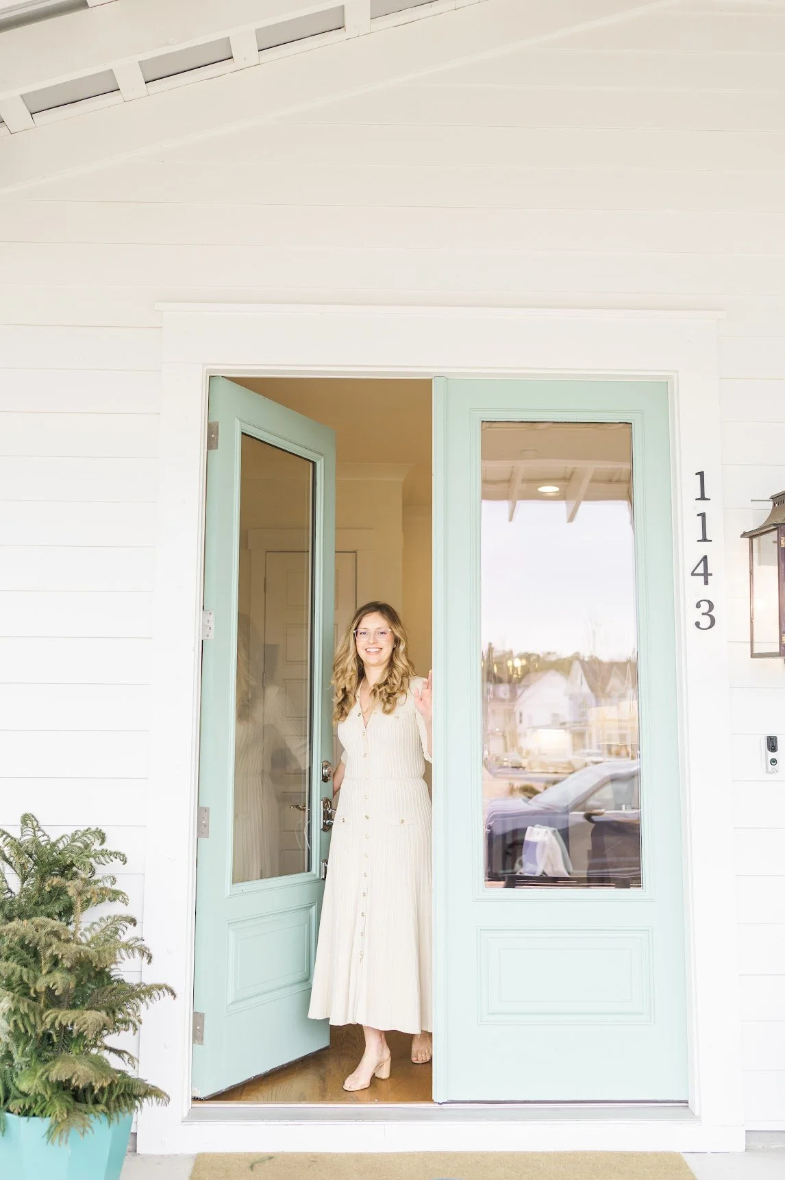 A woman with long blonde hair, glasses, and a cream-colored dress is smiling and standing in the doorway of a house, holding the door open. The house has white exterior walls, light blue front doors with glass panels, and house number 1143 displayed vertically.