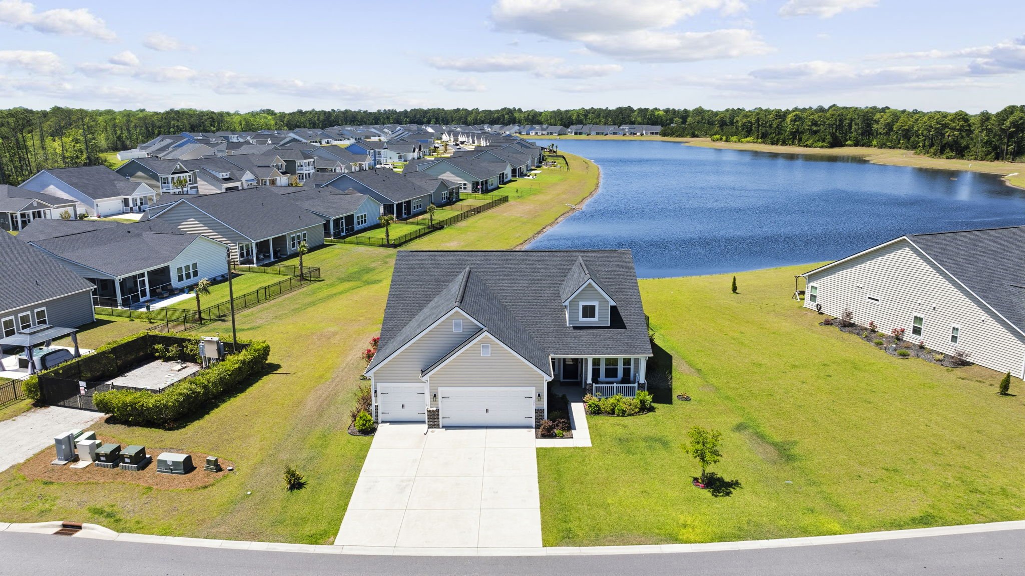 Aerial view of a suburban neighborhood with houses along a lake, surrounded by green trees under a partly cloudy sky.