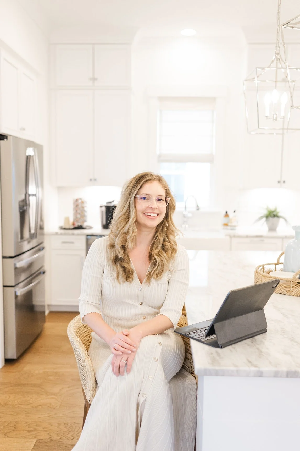 A woman with long wavy blonde hair, wearing glasses and a cream-colored dress, sitting at a kitchen counter with a tablet in front of her, smiling at the camera.