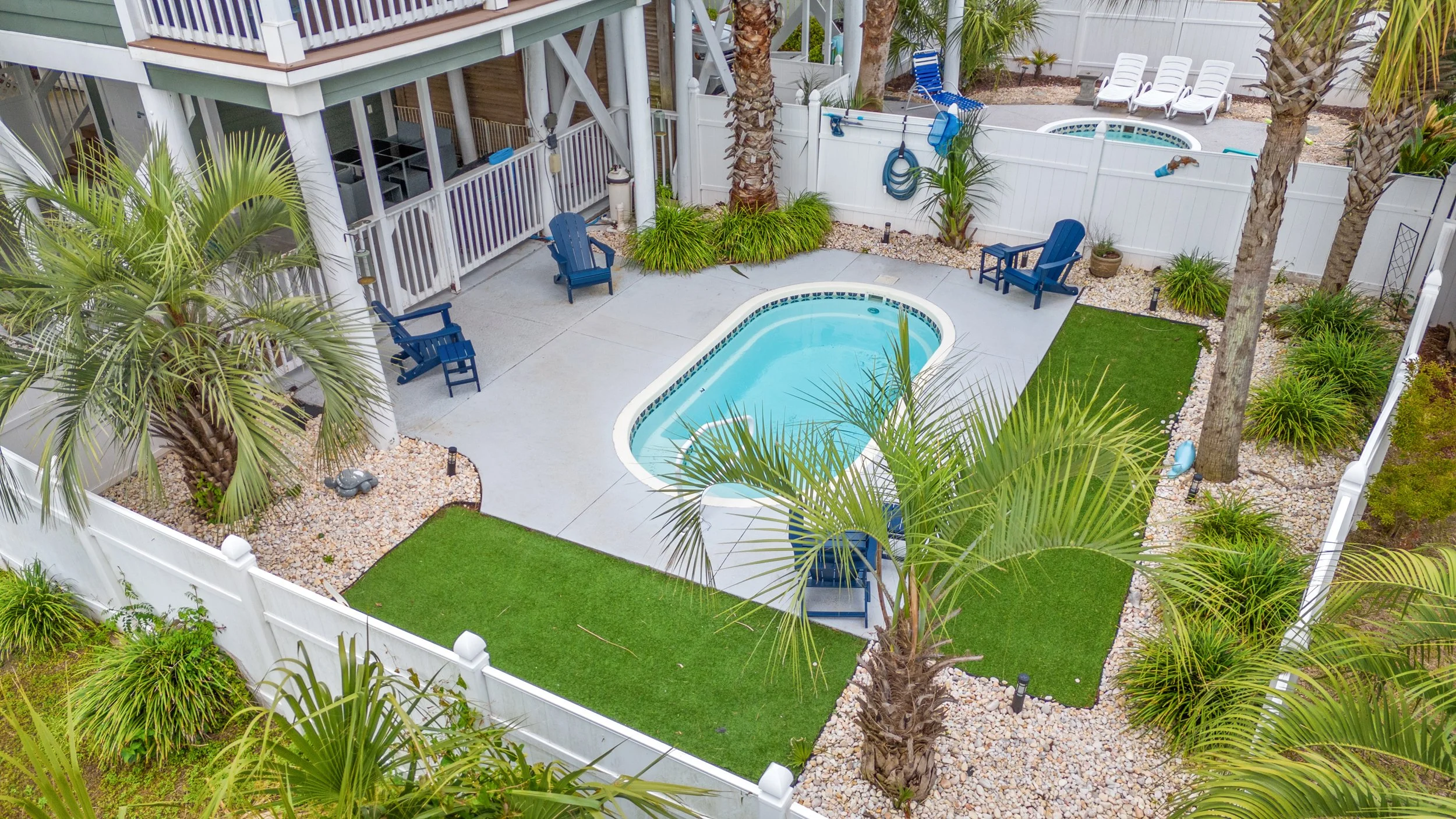 A backyard with a small swimming pool, outdoor seating, palm trees, and a white fence. There are several chairs around the pool area and a small lawn, with some decorative plants and gravel. A balcony top is visible at the top of the image.