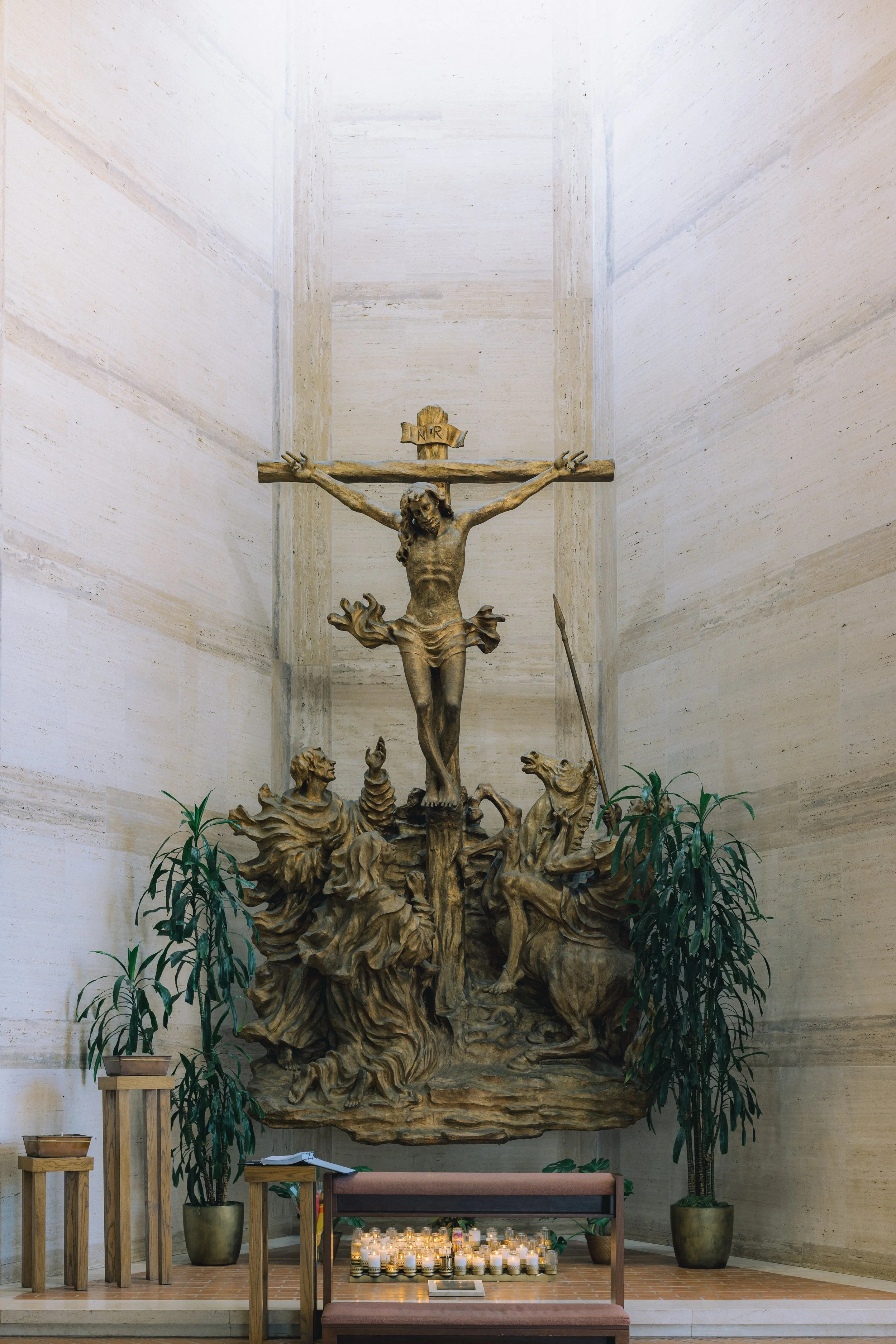 Religious altar with a large bronze crucifix of Jesus on wood, surrounded by two potted plants, candles on steps, and hymn books, inside a church.