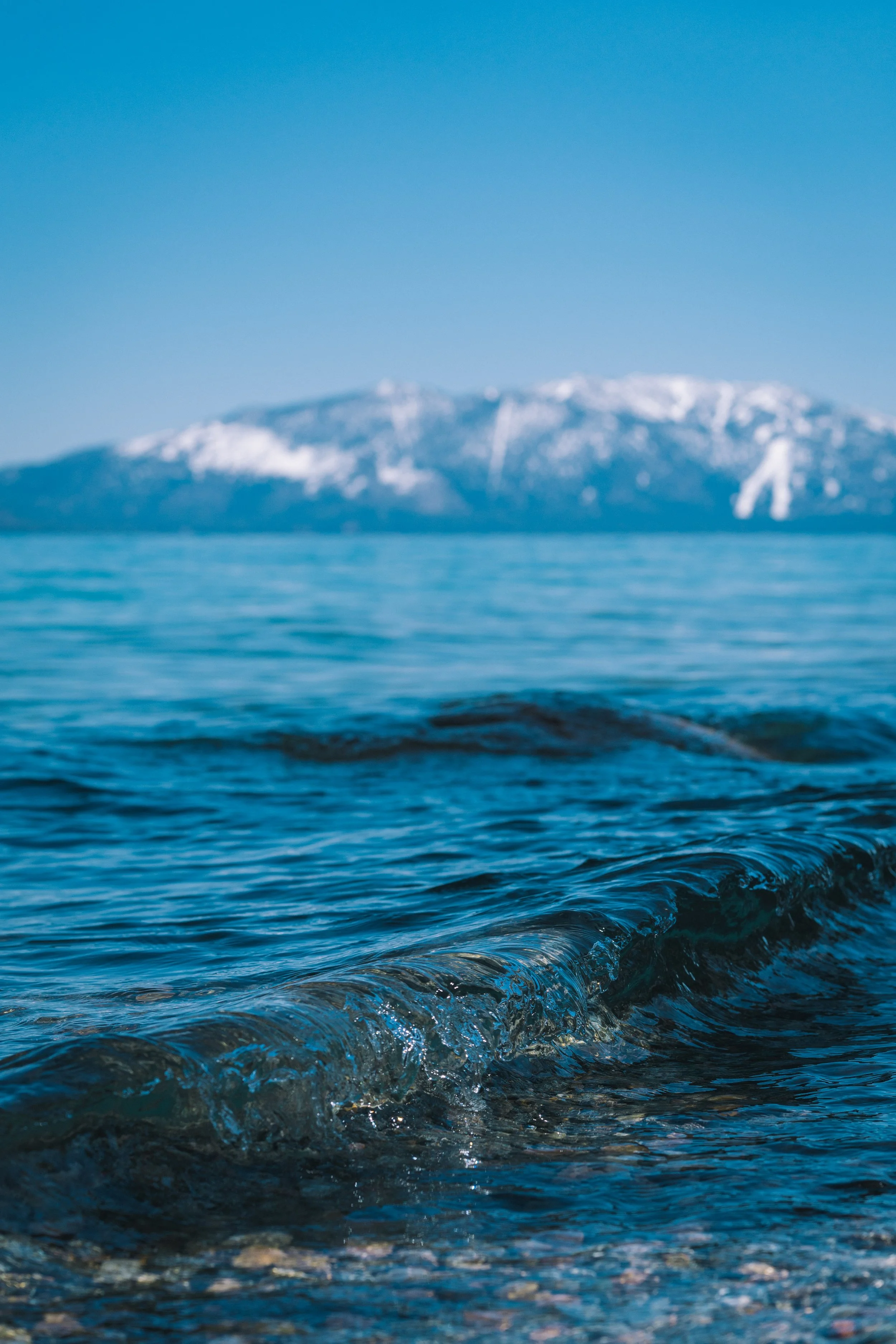 Close-up of ocean waves with a mountain range and snow-capped peaks in the background.