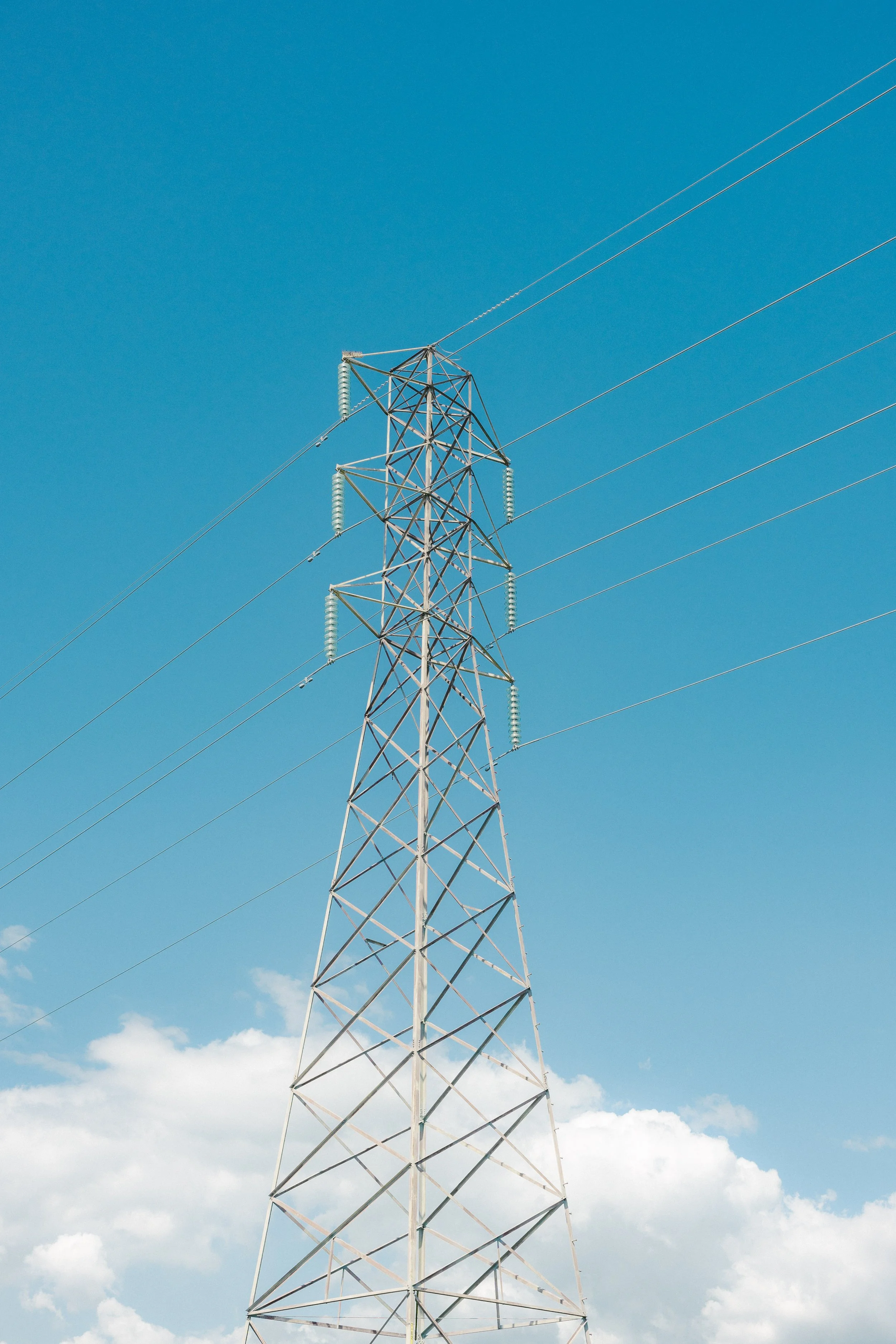 A tall metal electrical transmission tower with three power lines extending from it, set against a blue sky with scattered clouds.
