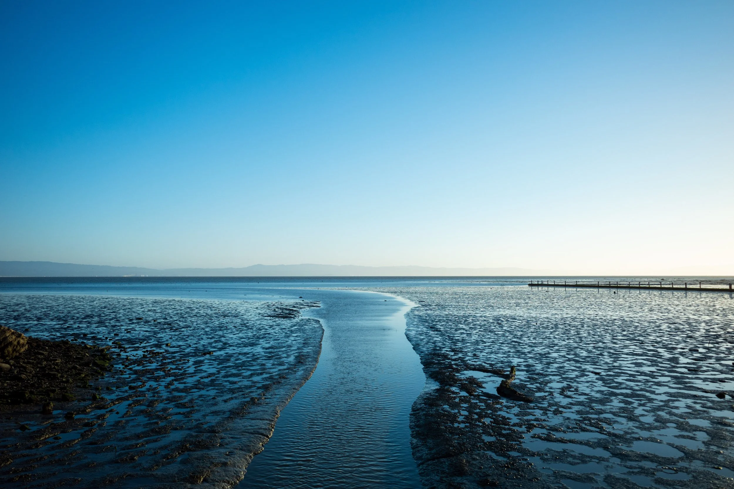 A calm and expansive river or bay during daytime, with a narrow waterway leading to the horizon, bordered by rocky shores and a distant mountain range under a clear blue sky.
