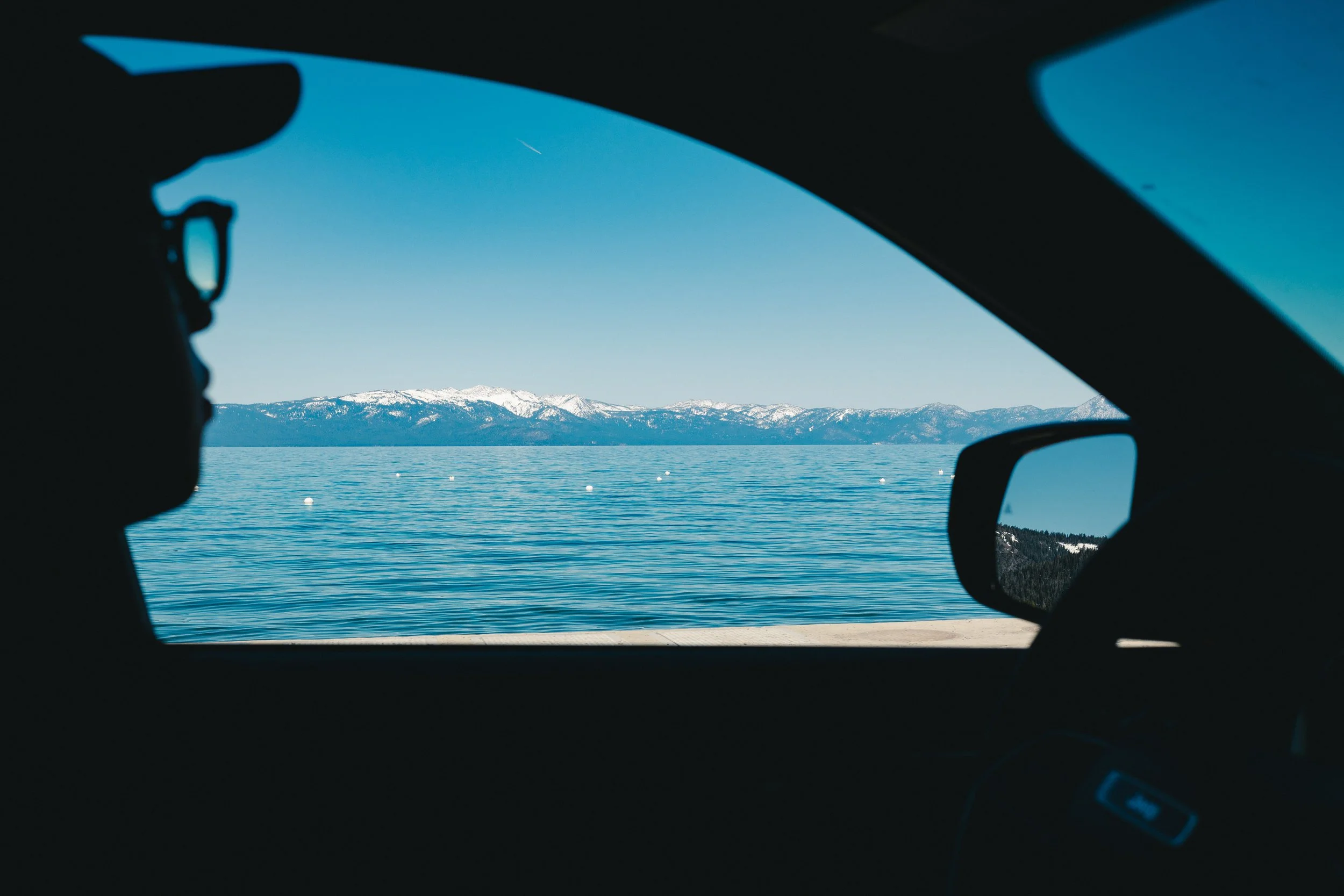 View from inside a car showing a lake with snow-capped mountains in the distance, seen through the windshield.