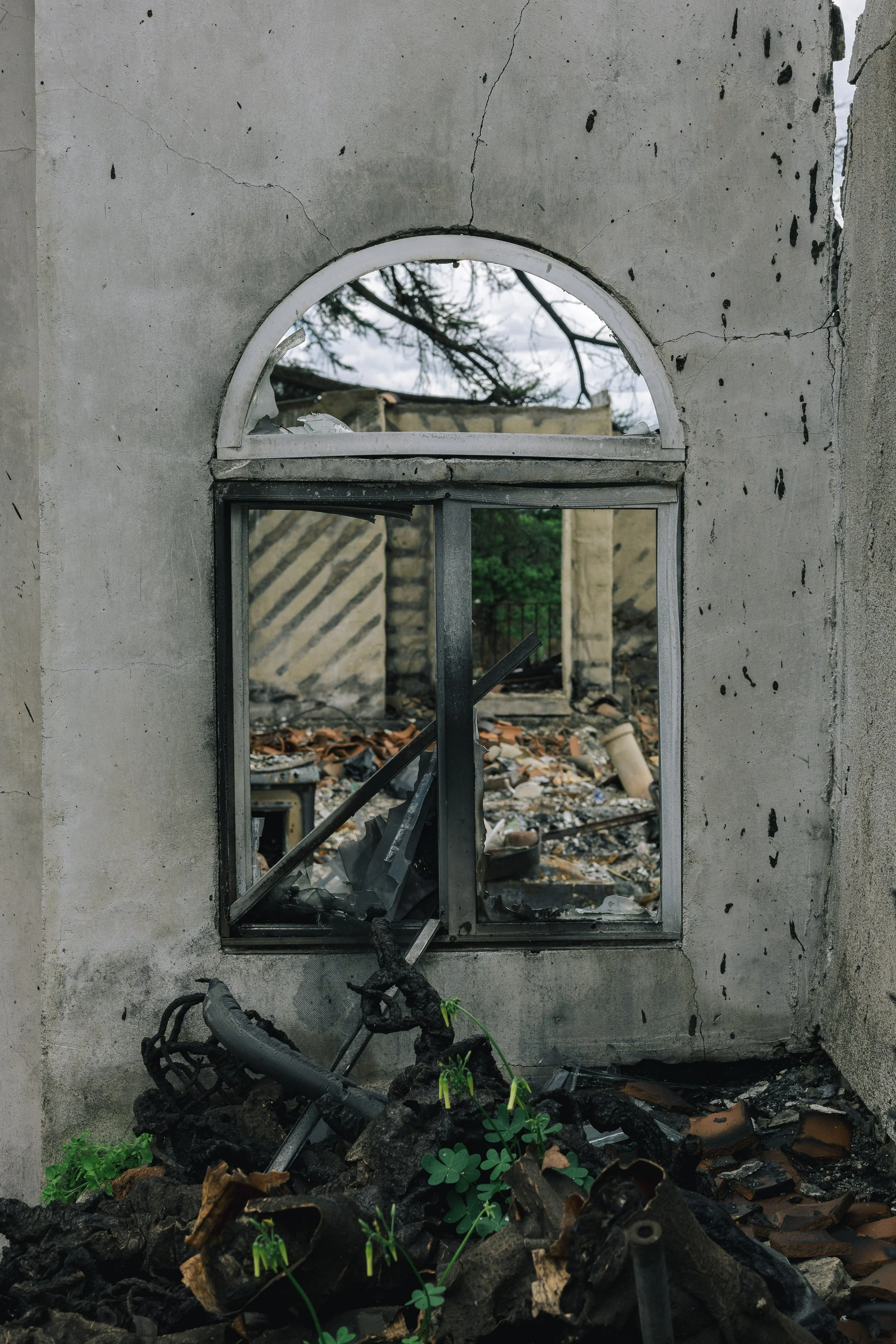 The remains of a burnt building with a window frame, and debris including burnt wood, metal, and ash. Inside, burnt rubble and broken structures are visible, with a fallen electrical wire and some small green plants growing at the base.