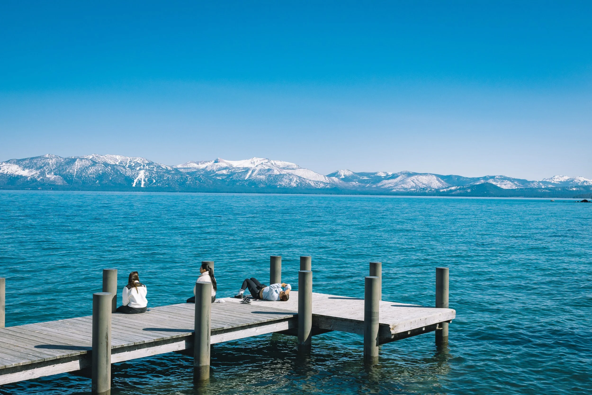 People relaxing on a wooden pier by a large body of water with snow-capped mountains in the background under a clear blue sky.