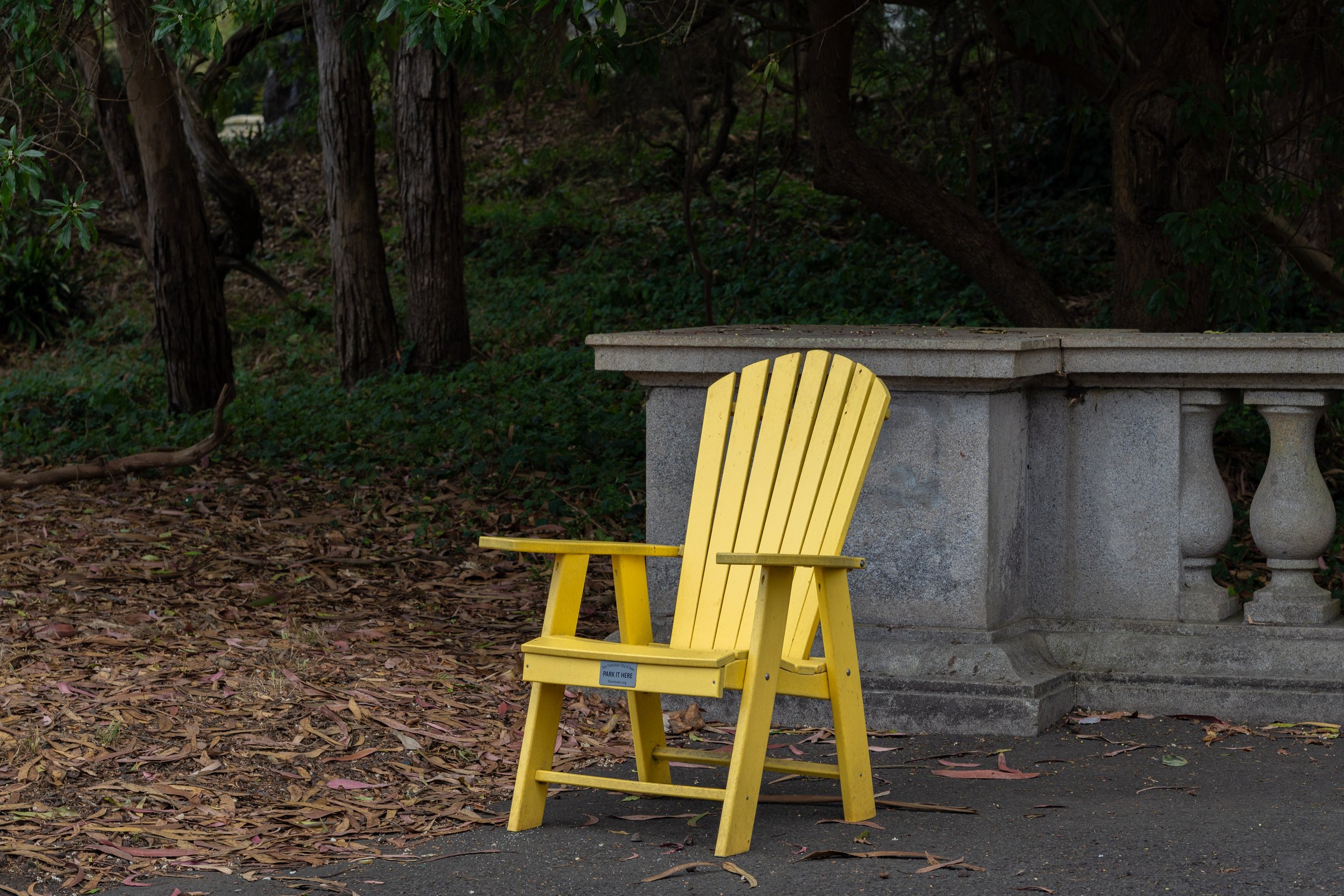 Yellow Adirondack chair in front of a stone or concrete garden bench, surrounded by trees and fallen leaves.