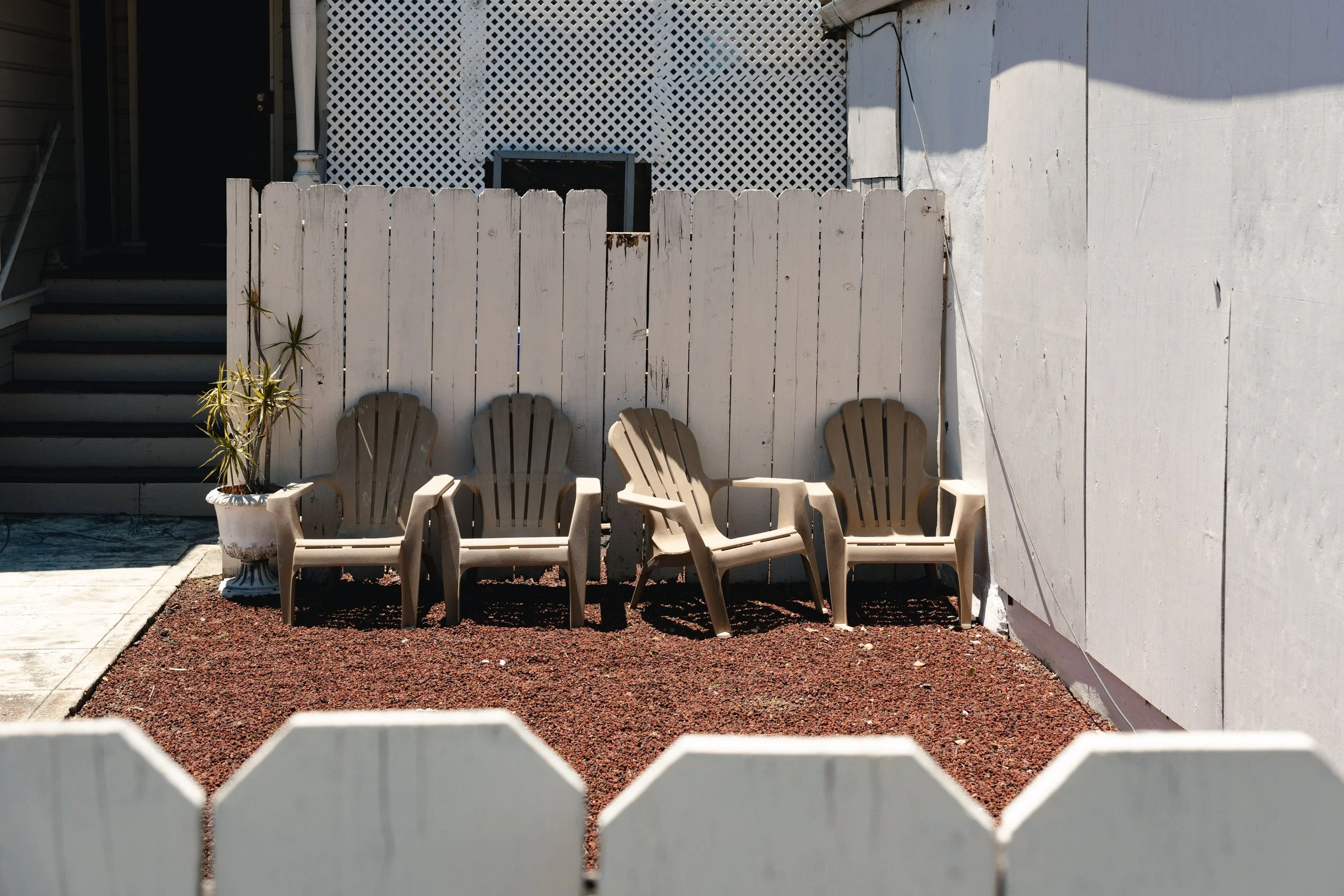 View of three plastic chairs and potted plant outside a white fenced yard, with white walls and lattice fence background, seen through a white picket fence in the foreground.