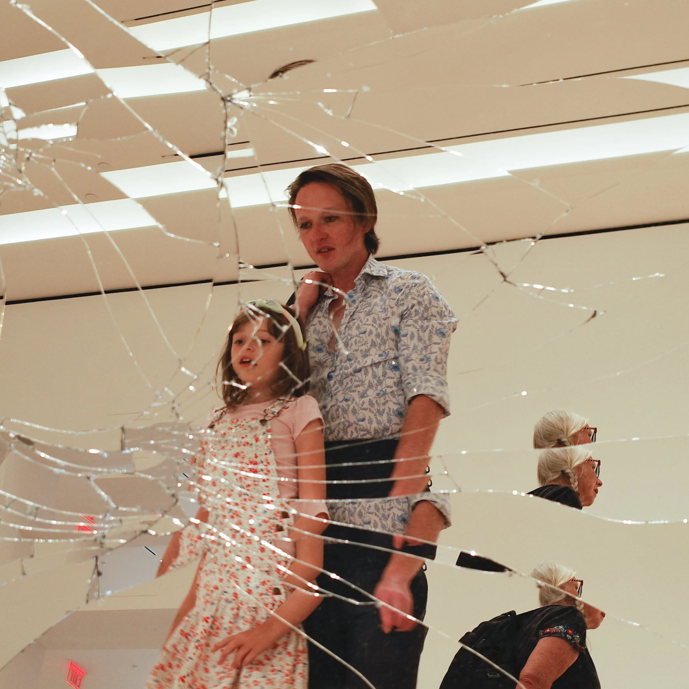 A woman and a young girl looking at a large mirror with a shattered glass pattern, reflecting the ceiling and people in an indoor setting.