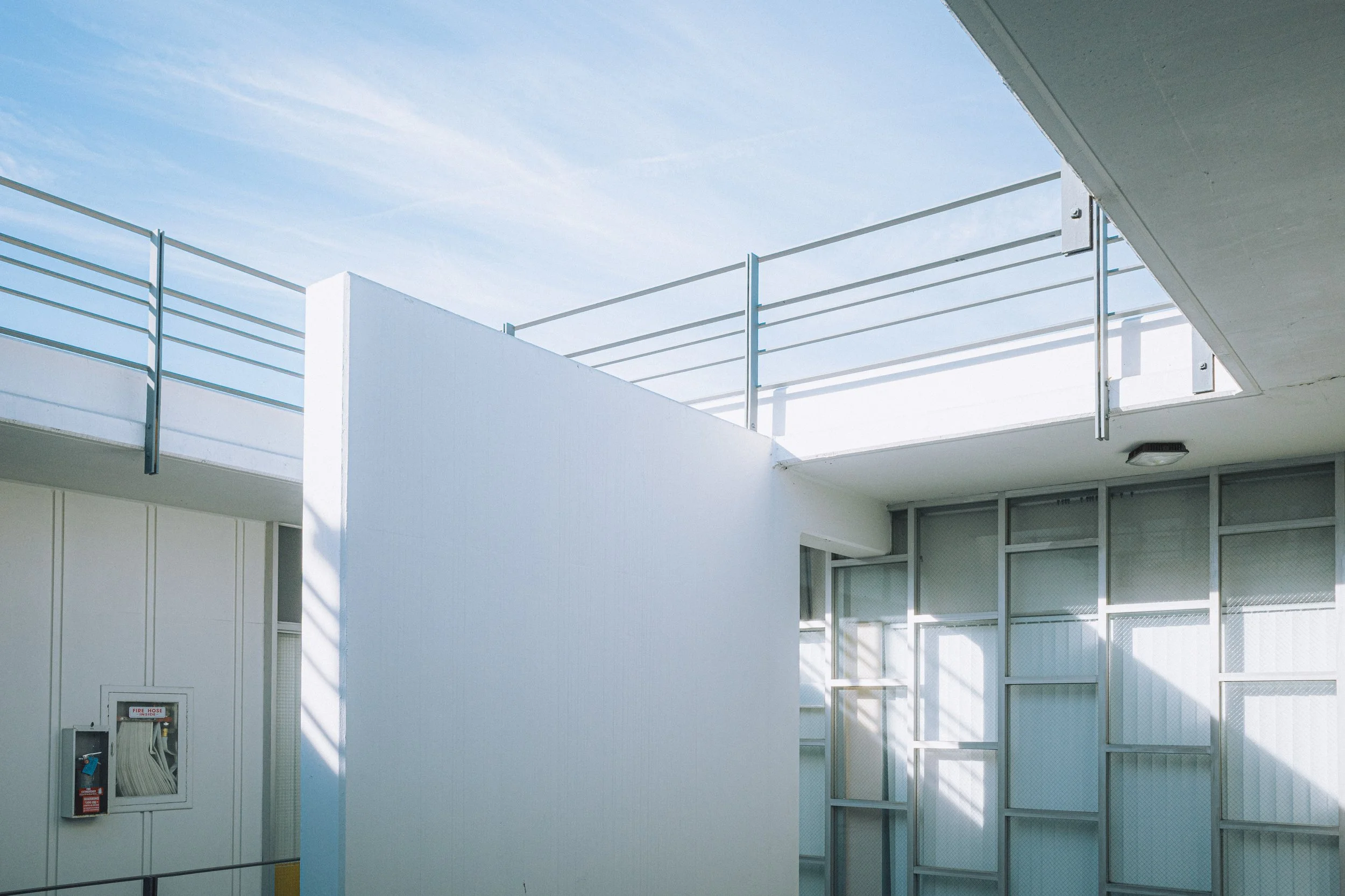 Photo of a modern building's exterior with white walls, glass windows, and a metal railing on the balcony, under a clear blue sky.