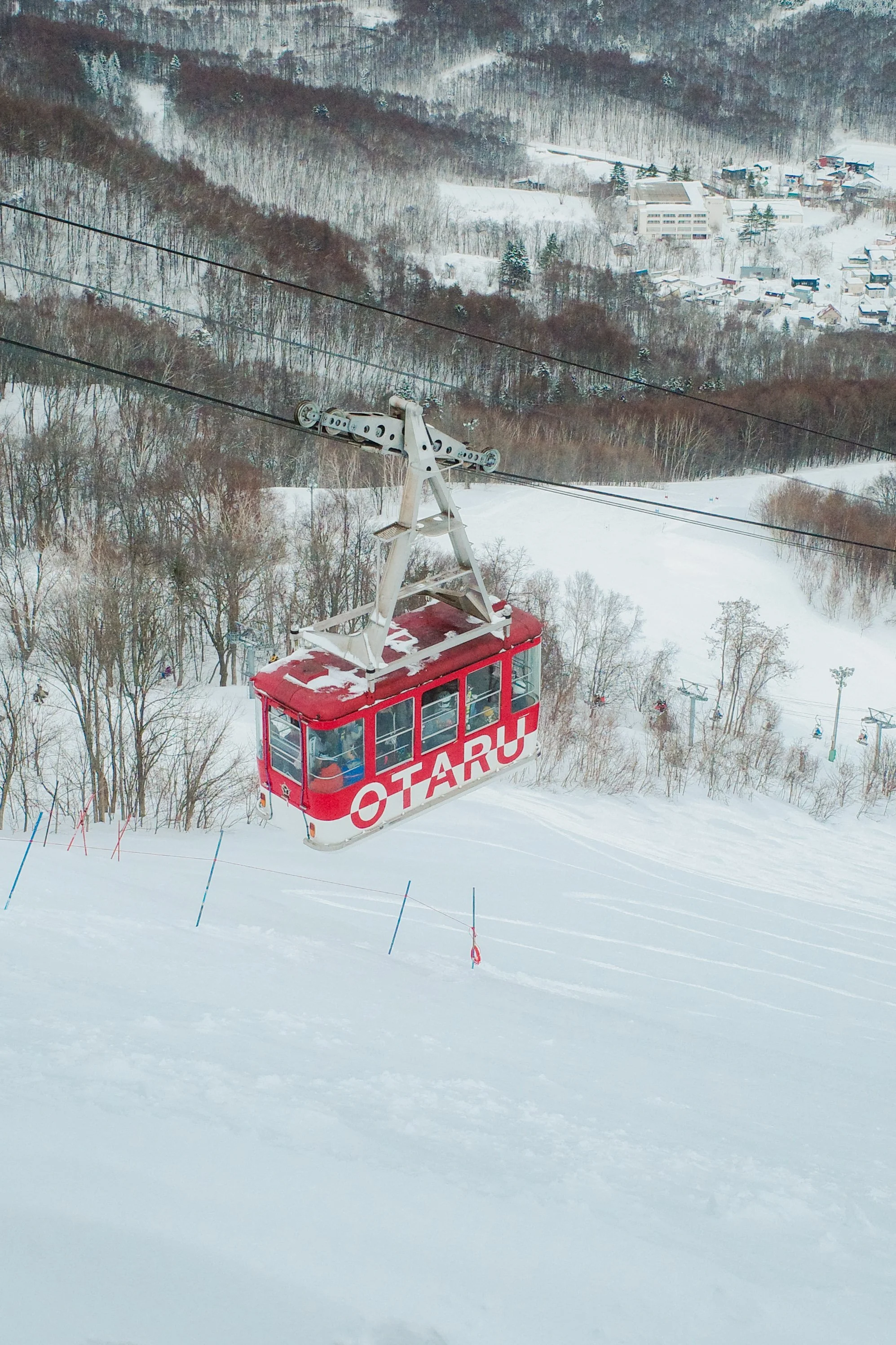 A red gondola lift named Otaru ascending above snowy ski slopes with snow-covered trees and a small town visible in the background during winter.