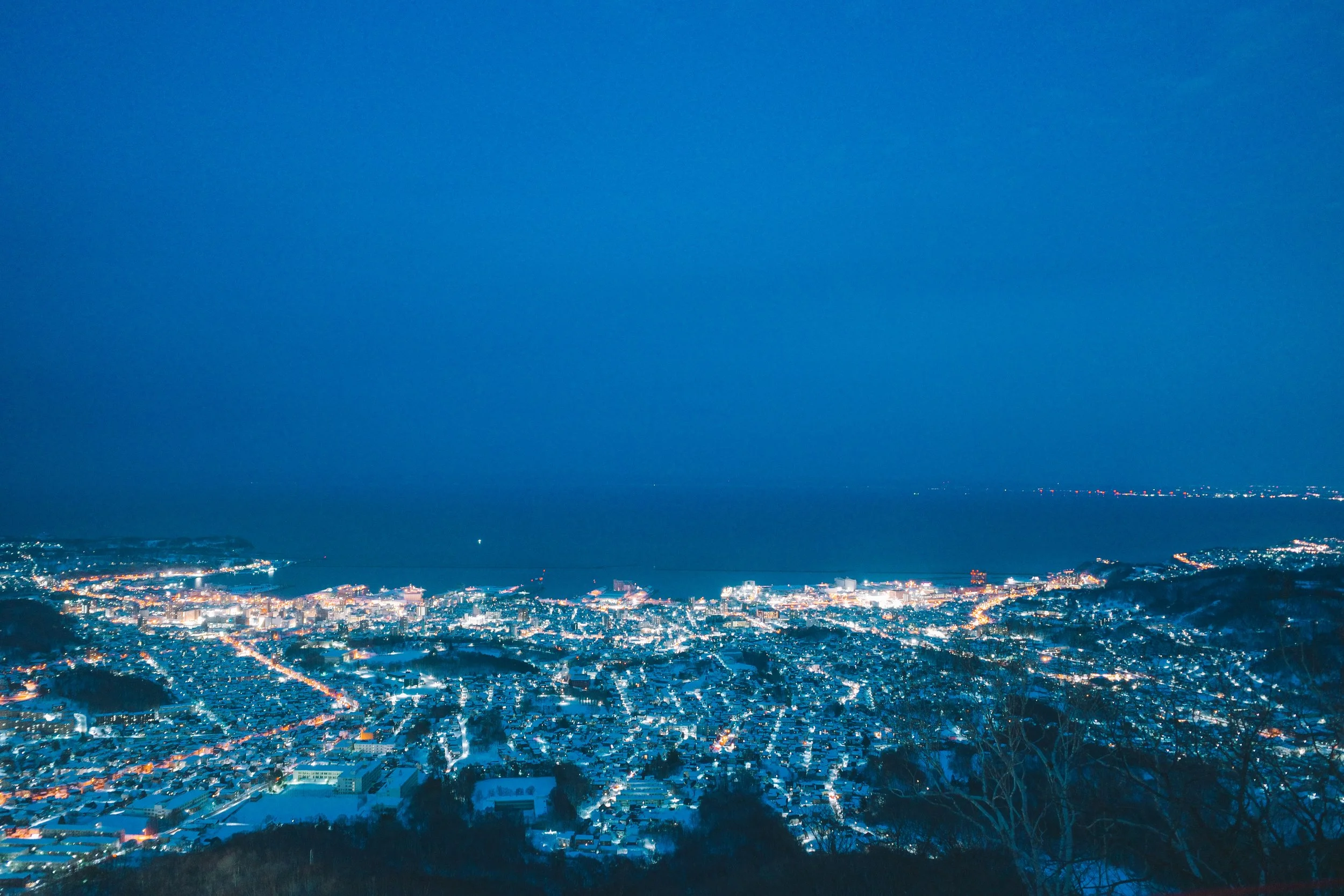 Aerial night view of a city with illuminated streets and buildings along a coastline, under a clear blue sky.